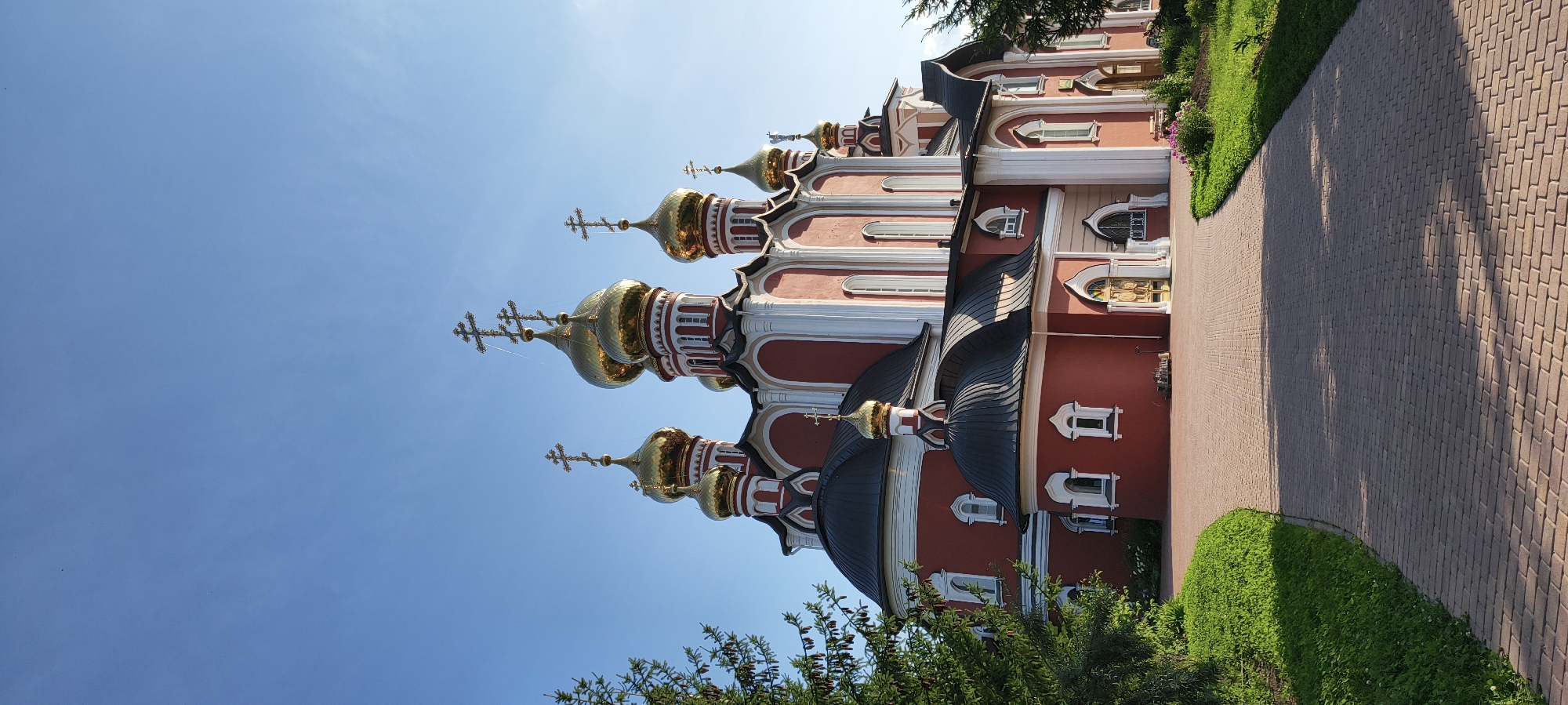 The image shows a bright, red Orthodox church with multiple golden domes, set against a clear blue sky. The church has white architectural details and is surrounded by a paved walkway and green grass. A tree with green foliage is visible on the left side of the image. The scene is bathed in sunlight, casting shadows on the ground.