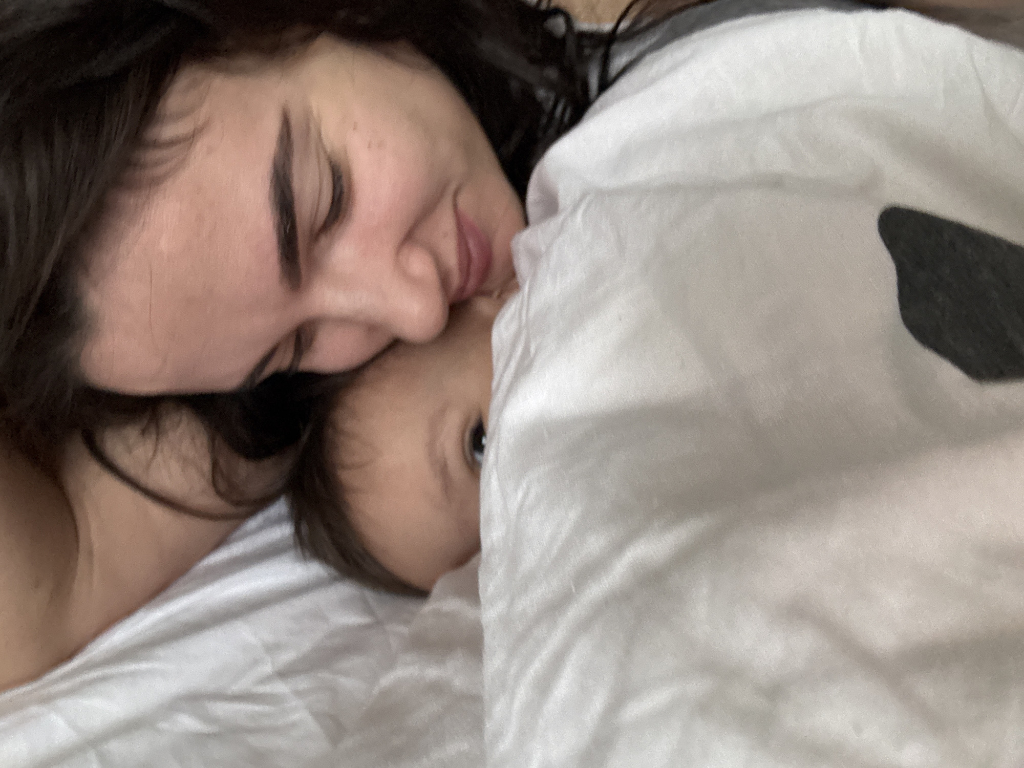 A woman lies on a white bed next to a baby partially covered by a white blanket with a black pattern. The woman has dark hair and is lying on her side with her eyes closed and a slight smile. The baby's head is visible near the woman's cheek, with one eye open and looking towards the viewer. The background is out of focus, providing little context about the location.