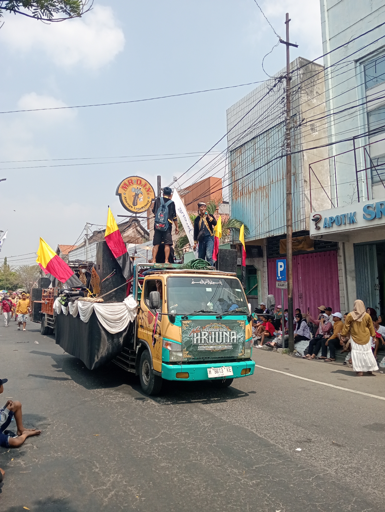This is a medium shot, eye-level, outdoor, daytime photograph taken in Demak, Indonesia. The scene depicts a parade or procession on a city street. A decorated truck, with a large platform and speakers, is the central focus. On the platform, three men are visible. Two are standing, and one is seated. One standing man is wearing a black backpack and shorts, and the other is wearing traditional attire and holding a microphone. Two flags, one yellow and red and the other black, are planted on the truck's platform. The truck is driving on a paved road. On the right side of the road, several people are seated, watching the procession. A building with the sign "APOTIK SRI" is visible in the background. The sky is partly cloudy.