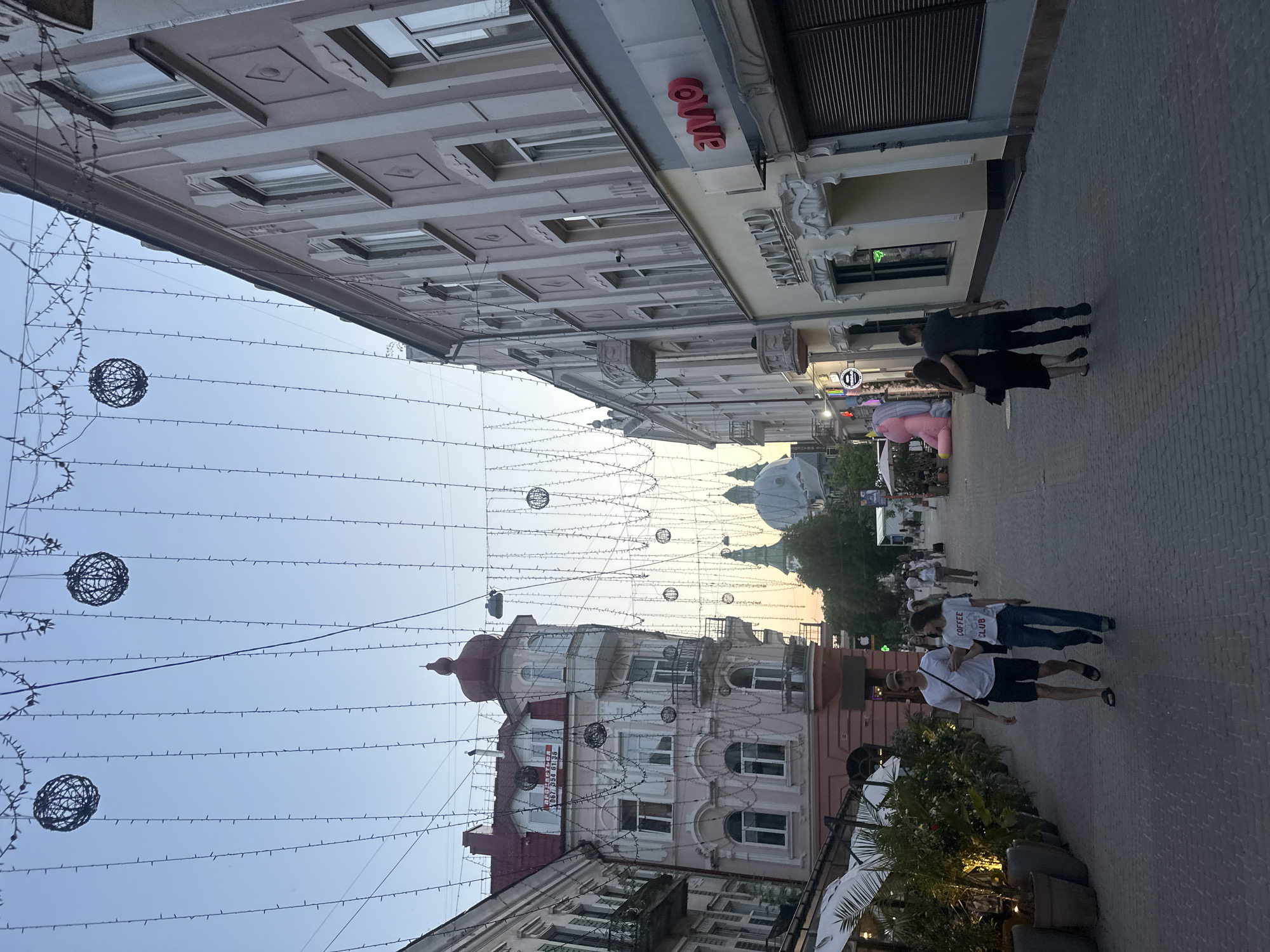 The image depicts a street scene in Ternopil, Ukraine, with buildings lining both sides of a cobblestone street. Overhead, strings of lights and decorative wire spheres are strung across the street. In the foreground, a couple walks away from the camera, with the woman wearing a t-shirt that says "COFFEE CLUB". Further down the street, another couple walks arm-in-arm. On the left side of the street, a building with a red roof and ornate architecture stands out. On the right side, a building has a prominent red logo that reads "anno". In the background, domed structures of churches are visible. The overall atmosphere suggests a pleasant evening stroll in a city center.