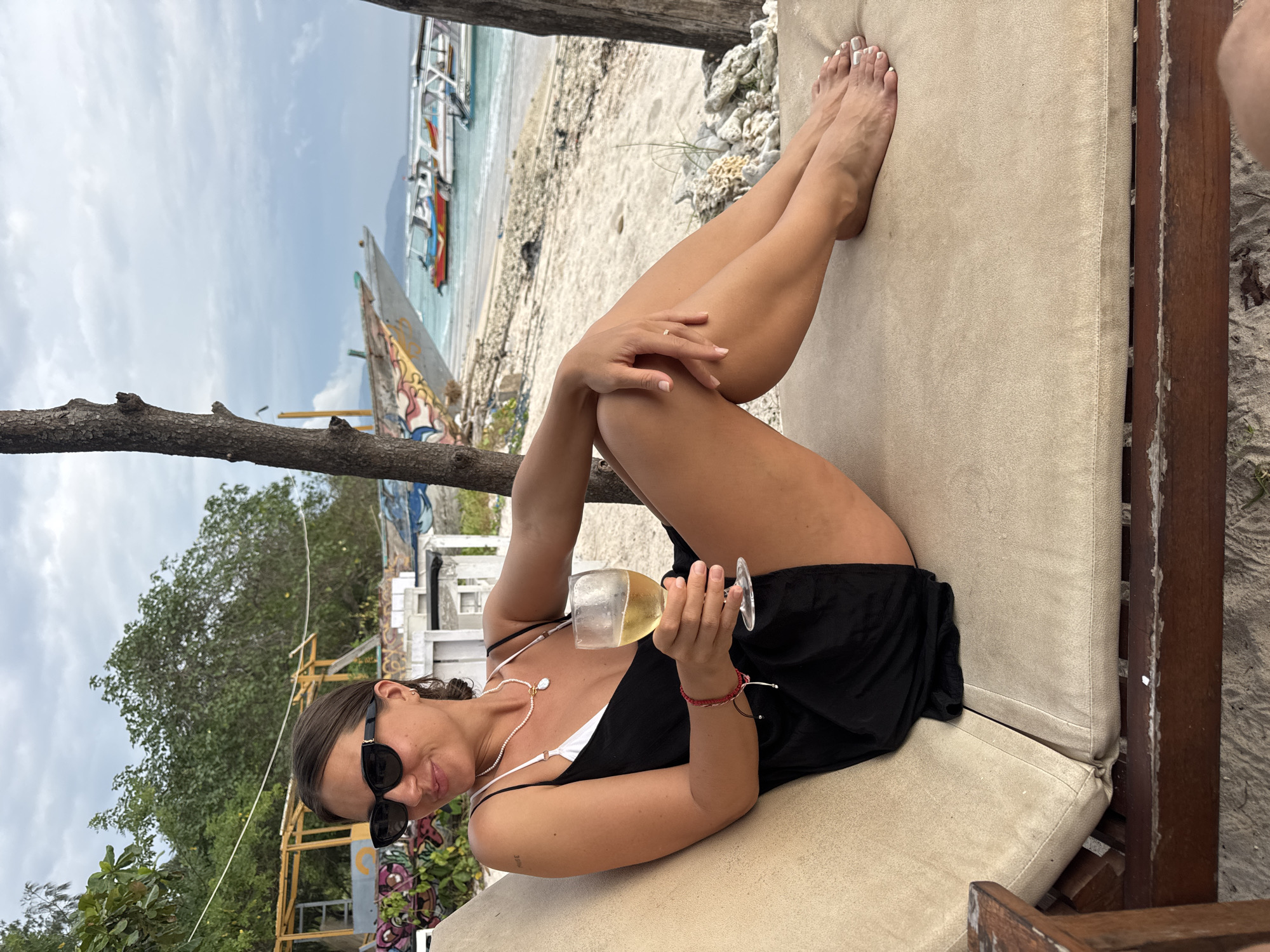 A woman is reclining on a beige cushioned lounge chair on a beach in Unknown, Indonesia. She is holding a wine glass filled with a clear liquid. She wears sunglasses, a black dress over a white top, and jewelry including a necklace, a bracelet, and a ring. The background includes trees, buildings, boats on the beach, sand, and the ocean. The sky is cloudy.