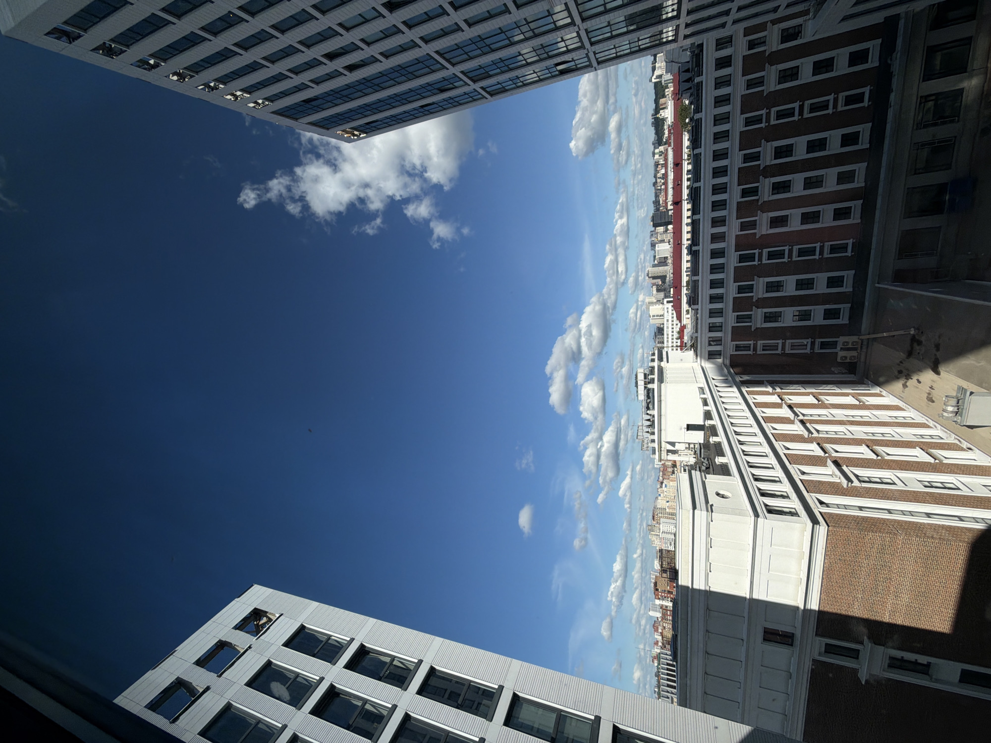 The image is an exterior shot, taken from a high vantage point looking upwards and outwards towards a cityscape under a bright blue sky with scattered white clouds. The perspective is tilted, making the buildings appear to lean.
In the foreground and extending across the top left of the frame is a modern building with a facade of horizontal grey panels and rows of dark-framed windows. To the right, and occupying the majority of the frame, is a longer, older building with a distinct red brick facade and white-trimmed windows arranged in a grid pattern. This building has a more ornate architectural style with white decorative elements and a prominent circular detail on one section. Further in the distance, a dense urban skyline of mixed-height buildings stretches towards the horizon.
There are no people visible. The scene depicts a quiet day in a city, likely mid-day given the bright sunlight casting shadows from the buildings. The weather appears clear and pleasant, with a few fluffy clouds dotting the expansive sky. The architecture suggests an urban setting with a mix of historical and contemporary styles. There is no visible text in the image. The overall impression is one of urban architecture viewed from above on a clear day in Ufa, Russia.