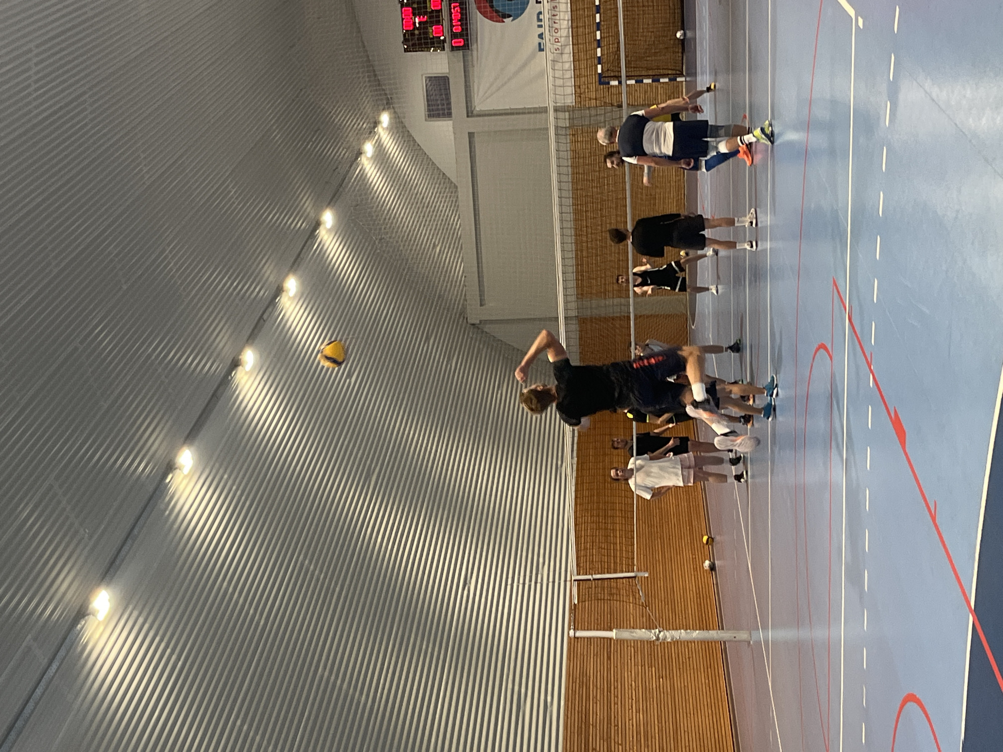The scene depicts a handball game or practice indoors, likely in Pobrdje, Montenegro based on the presence of a scoreboard with 'FAIR PLAY Sports' visible. Several people are present, with one individual in mid-air, appearing to spike or hit a yellow handball. Other players are positioned around the court. The location is a gymnasium with a blue playing surface, a net dividing the court, wood paneling, and a corrugated metal roof with overhead lighting. A scoreboard is visible in the background.