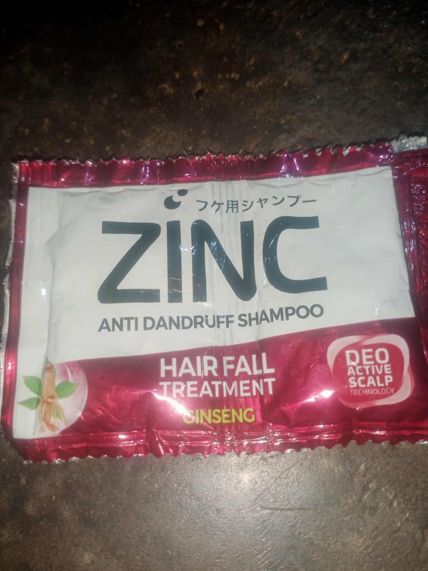 A close-up shot shows a sachet of "ZINC ANTI DANDRUFF SHAMPOO" with "HAIR FALL TREATMENT" and "GINSENG" text. The packaging is predominantly white and red, with some Japanese text at the top. A small illustration of ginseng root is visible on the left side of the sachet. A circular logo on the right side reads "DEO ACTIVE SCALP TECHNOLOGY." The sachet is placed on a dark, textured surface. The location of Nusa Agung, Indonesia is not depicted or implied in the image.