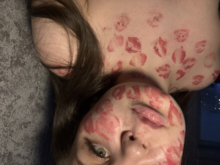 A person is lying down, covered in lipstick kisses all over their face, chest, and neck. They have long, dark hair and light eyes. The background appears to be a grey surface with a window revealing a cityscape at night. The person's expression is neutral.