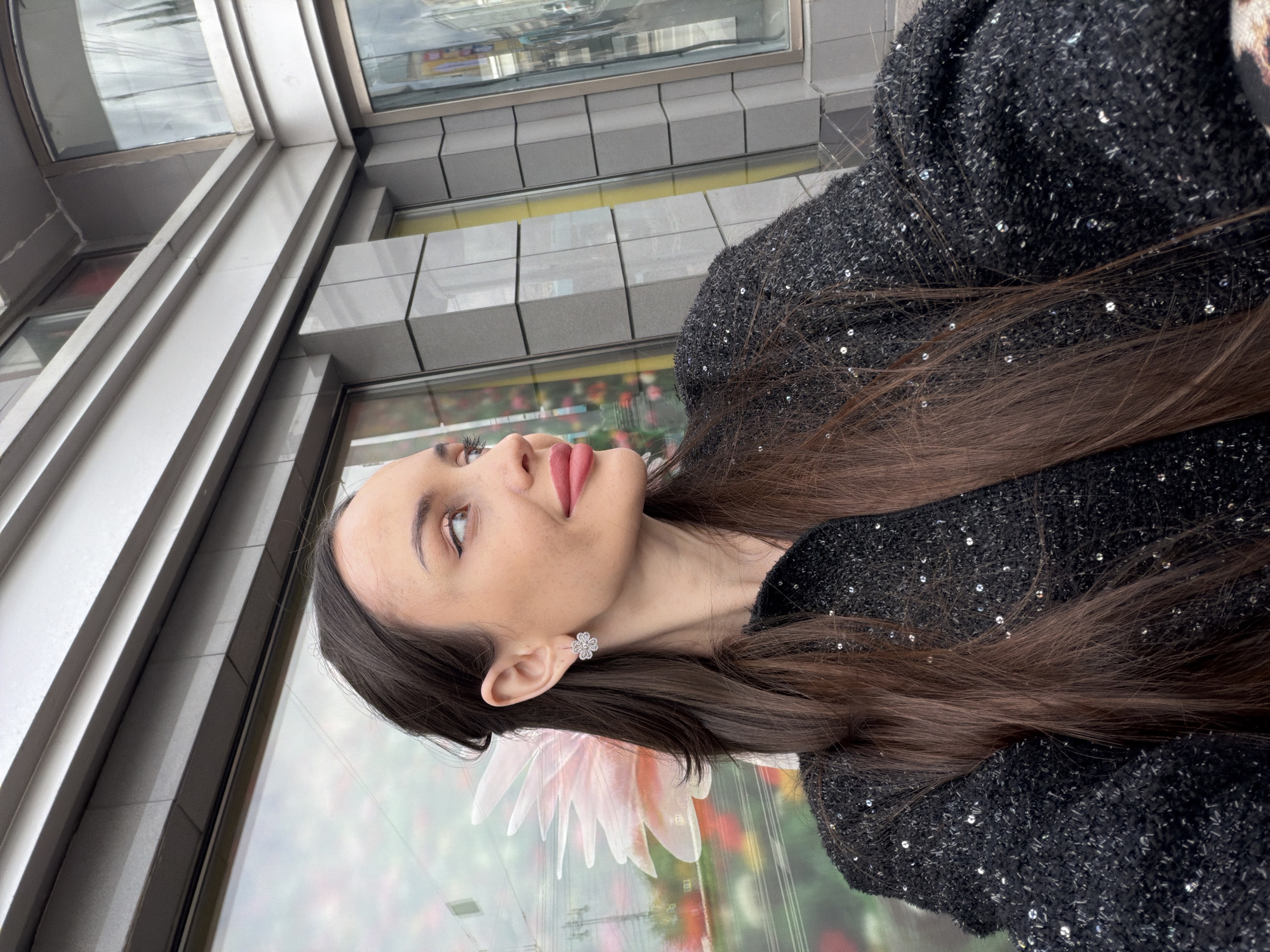 A woman is depicted in profile, looking upwards and to the left. She has long brown hair, fair skin, and is wearing an earring and a black jacket adorned with sequins. The background reveals an urban environment, specifically the city of Saint Petersburg, Russia. The image suggests an outdoor setting near a building with reflective glass windows. The surroundings may contain flowers, which can be seen in the reflections on the glass.