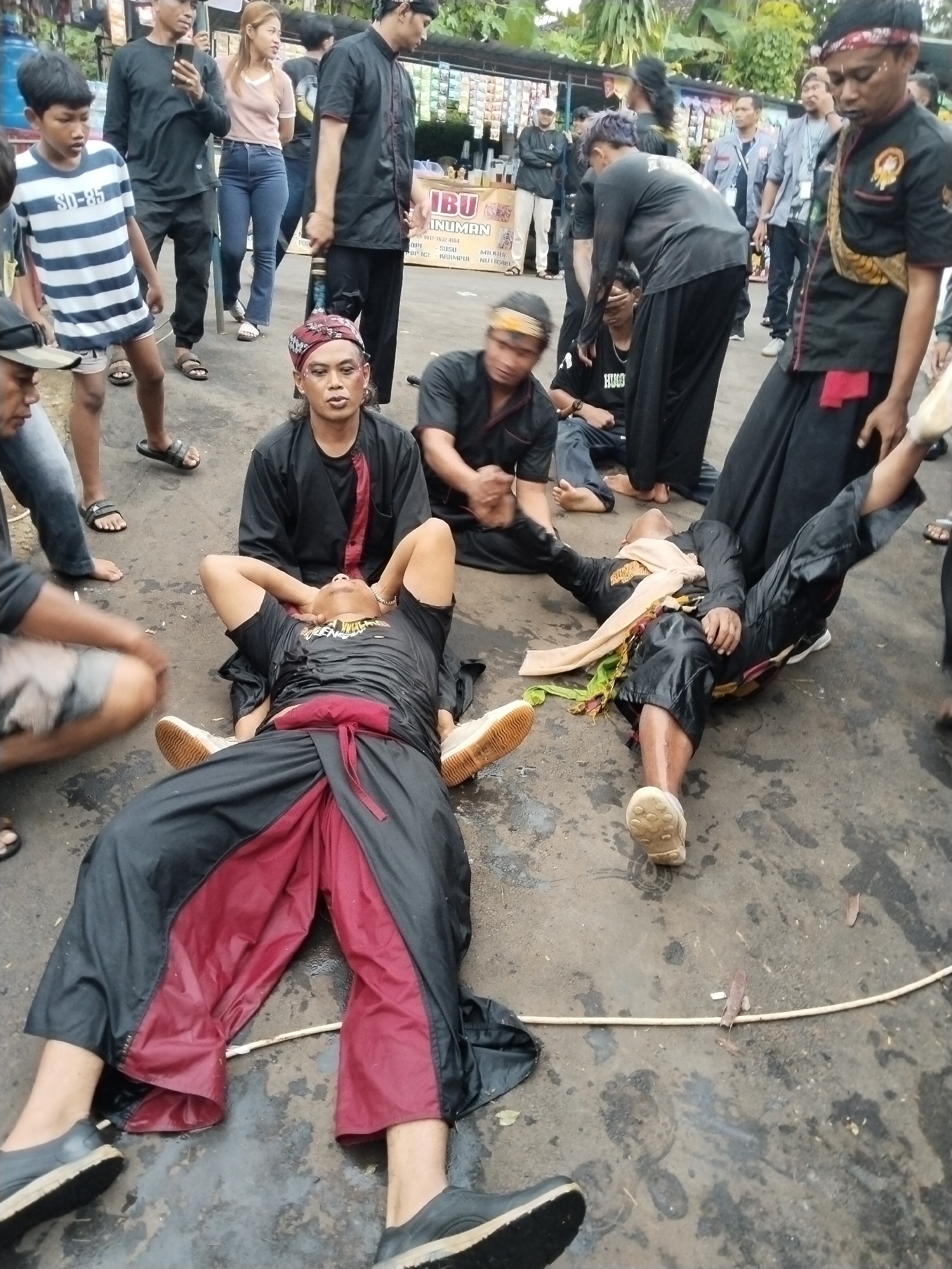 The image depicts a performance or ritual taking place in an outdoor space, likely in the city of Kuto, Indonesia. Several people are dressed in traditional black outfits with red accents, some wearing headbands or headdresses. Two individuals are lying on the ground, one with a scarf draped over them. Another man is seated nearby, seemingly attending to the person on the ground.
Several other people are standing around observing the scene. In the background, there is a storefront or stall selling beverages. The ground appears to be a paved surface, possibly a road or courtyard. The scene conveys a sense of cultural or spiritual significance.