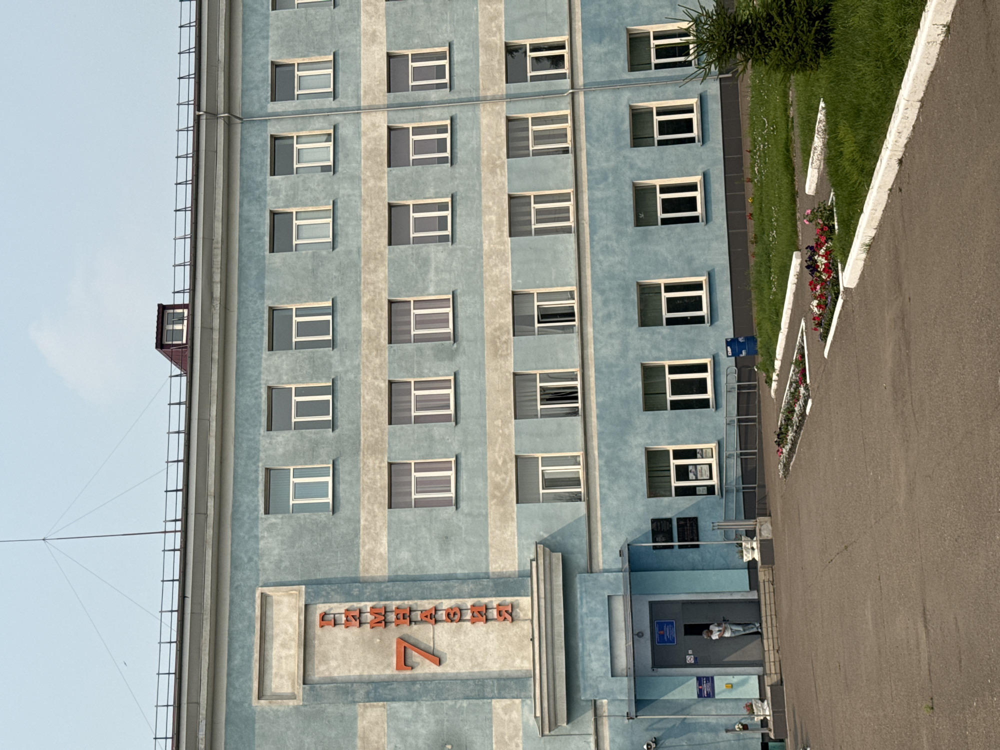 The image shows the exterior of a building, likely a school or administrative building, with light blue and beige colored walls. The building has multiple windows and signage in Russian. A prominent red sign on the left side of the building reads "7 Гимназия", indicating "Gymnasium 7". The building appears to be a multi-story structure. In front of the building, there is a paved area, possibly a courtyard or parking lot, with some landscaping including flower beds and a small evergreen tree. A single person, a woman wearing a white shirt and blue pants, is standing in an open doorway on the ground floor. The sky is clear with a hint of haze. The overall scene suggests an outdoor view of a public building in a city. While the specific location within Krasnoyarsk is not provided, the visual cues, like the architectural style and Cyrillic signage, are consistent with a Russian urban environment.