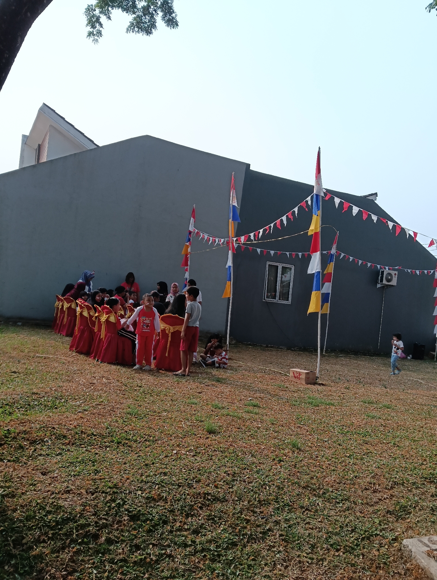 In Parung Panjang, Indonesia, a group of children, primarily young girls dressed in vibrant red attire, are gathered outdoors in front of a modern, gray-walled building. The children are seated on chairs adorned with gold sashes, suggesting a formal or ceremonial occasion. Several adult women, some wearing headscarves, are also present, appearing to supervise or participate in the event. The environment is characterized by a lawn area with patchy grass and dry leaves. The building's facade features a single window and an air conditioning unit. Decorative flags in red, white, yellow, and blue are strung across the scene, adding a festive atmosphere. A few children are standing and interacting, while others are seated on the ground. A small, light-colored box with the word "Vit" printed on it is visible on the grass. The sky is clear and bright, indicating daylight.