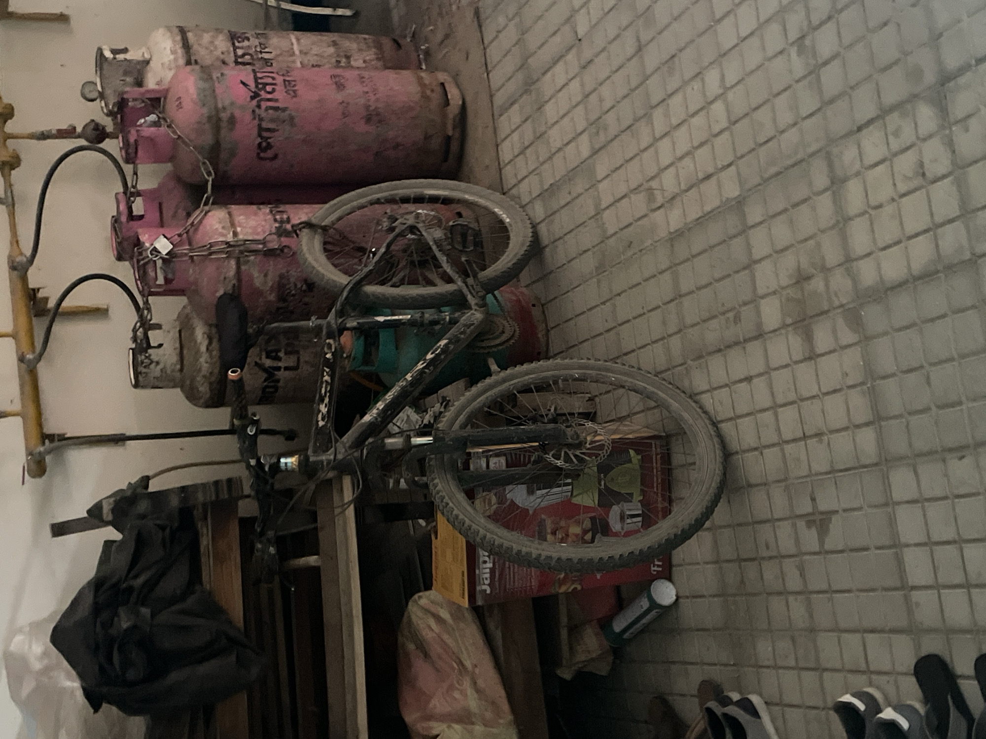The image depicts a cluttered indoor space in Dhaka, Bangladesh, likely a storeroom or utility area. In the foreground, a black mountain bike rests horizontally, partially obscuring a stack of five red gas cylinders chained together. A black plastic bag and various objects including an unopened box marked “Jaipur” and a piece of cloth appear near the base of the stack. The floor is tiled with small, square, gray tiles. The scene is illuminated by overhead light.
