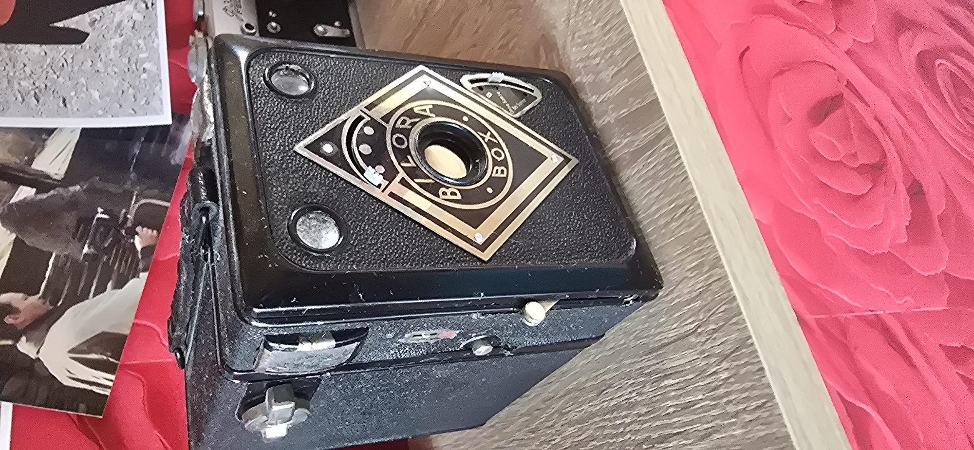 A black Bilora Box camera sits on a wooden surface in the foreground, angled slightly to the right. The camera has a textured body and a metallic diamond-shaped emblem on the front that reads "BILORA BOX" in gold lettering. Two circular viewing lenses are positioned above the emblem. A worn, black strap is attached to the top of the camera. To the left of the camera, there is a silver-colored knob and a latch mechanism. In the background, a photograph is partially visible, showing two individuals. One individual, on the left, is wearing a light-colored jacket over a darker garment and is holding a camera. The second individual, to the right, is mostly obscured. The background is a blurry mix of textures and colors. The scene appears to be indoors, likely on a shelf or table, with a bright pink, rose-patterned fabric in the lower portion of the image. The location is not identifiable from the image itself, and therefore, its presence in Gostagayevskaya, Russia cannot be confirmed.