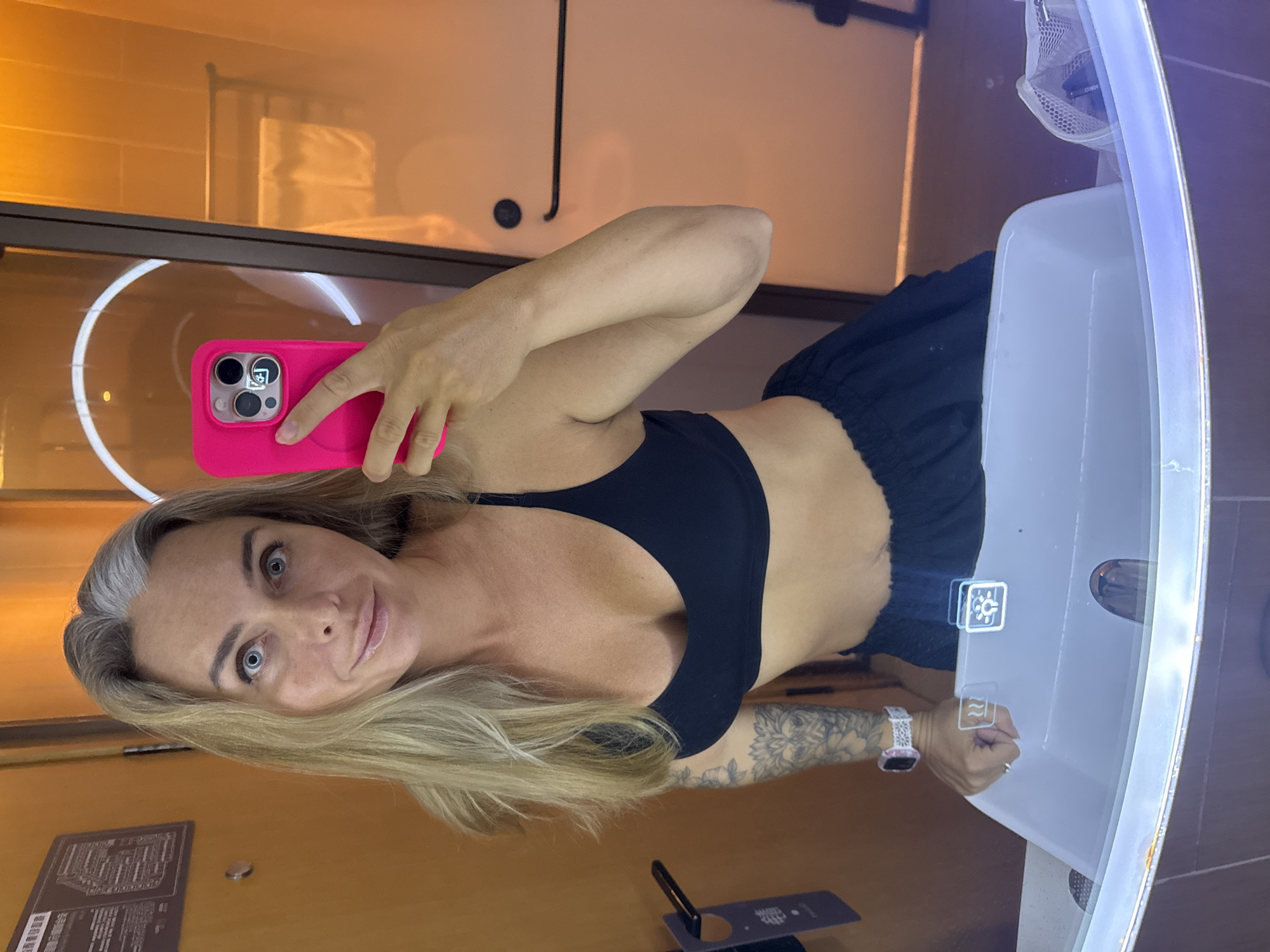 An adult Caucasian female with blonde hair and grey streaks, wearing a black sports bra and dark blue shorts, takes a selfie in a modern hotel bathroom. She is holding a pink smartphone with her right hand. Her left arm is visible, displaying tattoos. A white smart watch with a decorative band is worn on her left wrist. The bathroom features a large, illuminated sink and vanity with a mirror that reflects the scene. Behind her, a towel rack with a white towel and a circular light fixture are visible. The wall behind her appears to be wood-paneled with orange lighting, creating a warm ambiance. The image does not provide any specific clues or indicators that would allow for its localization to Shanghai, China.