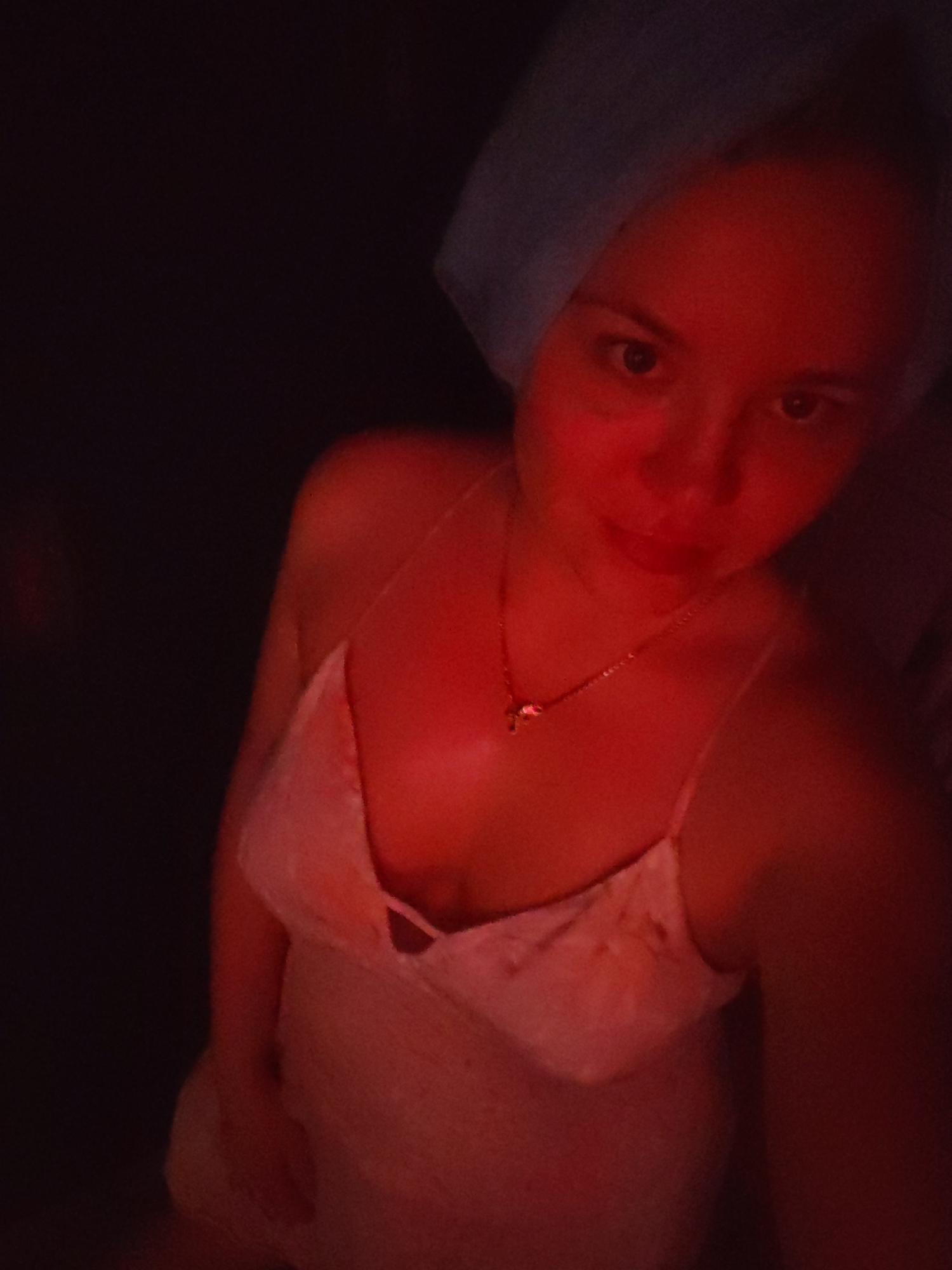 The image depicts a close-up, over-the-shoulder shot of a young Caucasian woman wearing a light pink slip and a blue headscarf. She is illuminated by red lighting, which casts a warm glow on her skin. Her eyes are open and she is looking towards the camera. A delicate gold necklace with a small pendant is visible around her neck. The background is dark and out of focus. The scene appears to be an indoor setting, possibly a bathroom or bedroom, given the headscarf and attire. There is no information in the image to suggest a specific location in Izhevsk, Russia.
