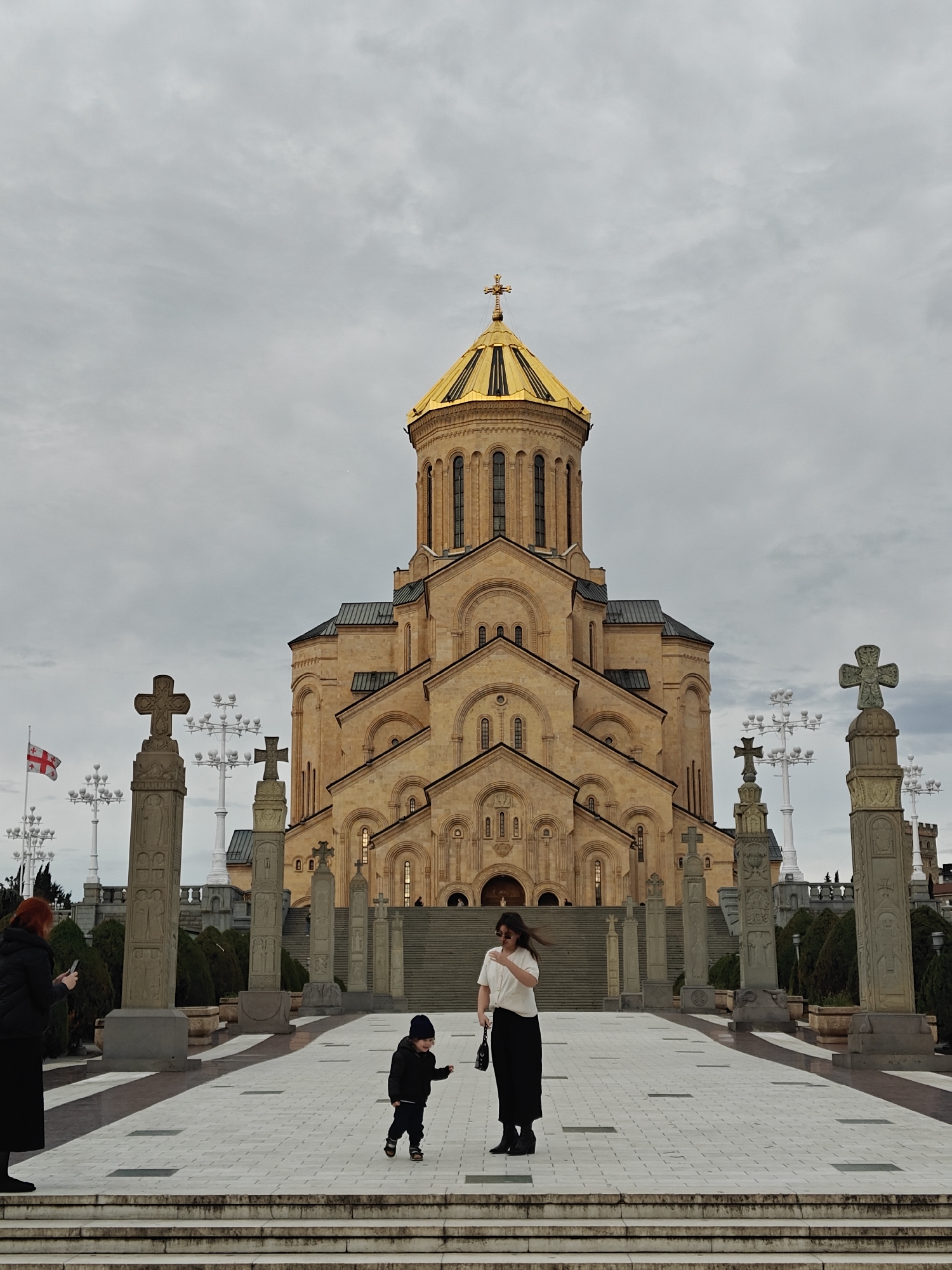 The image features the Holy Trinity Cathedral of Tbilisi, Georgia, under an overcast sky. The grand cathedral, built from light-colored stone, dominates the background with its complex, multi-tiered structure, crowned by a prominent golden dome and a golden cross at its apex.
A wide, paved plaza with a distinct tiled pattern leads up to the cathedral, incorporating several broad steps. This expansive area is flanked by numerous tall, intricately carved stone cross monuments (steles) arranged in rows. White, multi-globular lamp posts are also visible throughout the plaza. On the far left, a Georgian flag, white with a large red cross and four smaller red crosses in its quadrants, flies from a flagpole. Green shrubbery lines the edges of the plaza.
Three individuals are present in the foreground and midground. On the far left, a woman with reddish-brown hair, dressed in a black puffer jacket and a long black skirt, is holding a smartphone, seemingly taking a picture of the cathedral. In the center, a small child, a toddler wearing a black beanie and jacket, is actively walking on the tiled path. To the child's right stands another woman with dark hair, wearing a white short-sleeved top, a long black skirt, and sunglasses perched on her head; she carries a black handbag and her hair appears to be slightly windblown.
The sky is entirely overcast with light gray clouds, indicating an absence of direct sunlight and likely a cool, mild day. The scene captures visitors exploring and admiring the significant architectural landmark. No discernible text is visible on any of the structures or objects.