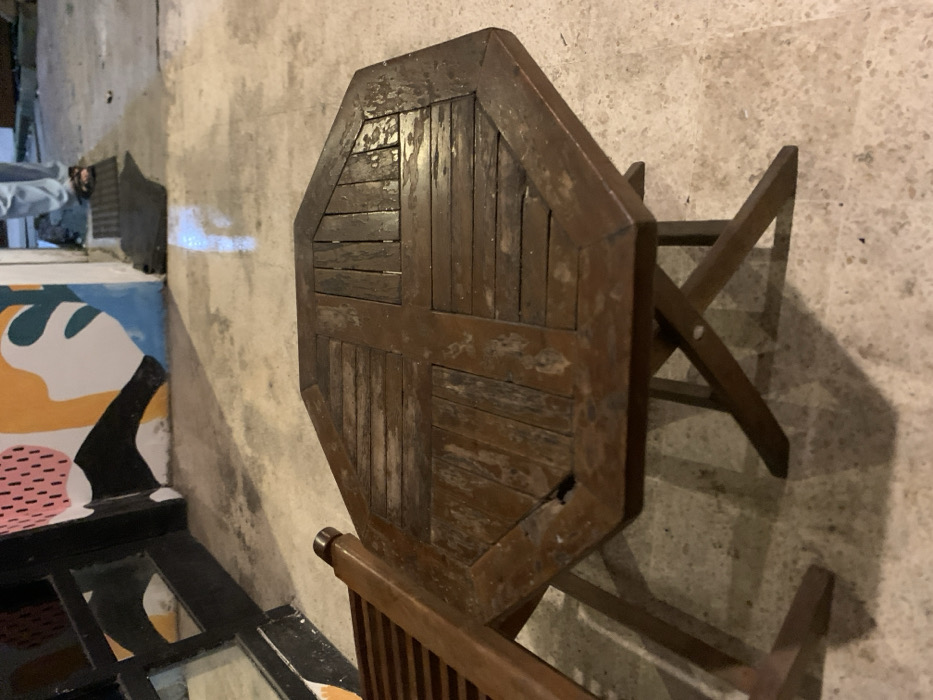 The image shows a wooden octagonal table with a wooden chair next to it. The table is made of dark brown wood with visible wood grain and a slightly distressed finish. The legs of the table are visible, and they are made of a similar wood to the top. The floor is a light gray concrete, and there is a white wall with a blue and yellow painted design in the background. The scene is located in Probolinggo, Indonesia, at the coordinates -7.751 latitude and 113.213 longitude. The image appears to have been taken indoors, possibly a cafe or restaurant.