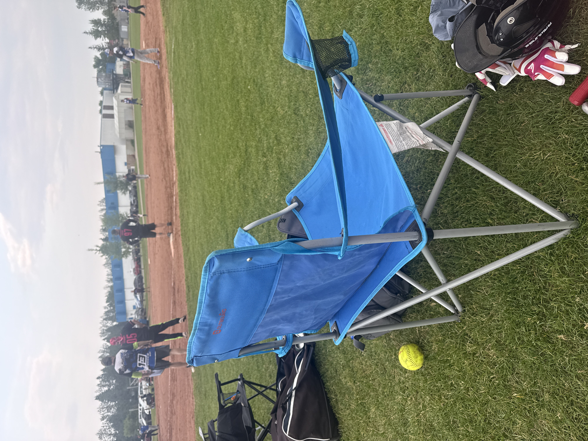 An outdoor daytime medium shot shows a bright blue camping chair positioned in the foreground on a grassy field, facing away from the camera. The chair has a mesh cup holder on its right arm. To the left of the chair, on the grass, rests a yellow softball. Behind the chair, several other camping chairs are visible, one dark-colored and folded. A large black duffel bag lies on its side next to the chairs. In the background, a dirt baseball field is in play with several individuals in uniform on the field and spectators in the distance. A blue and white building with a grey roof sits behind the baseball field. Trees and parked vehicles are visible beyond the field. The sky is overcast with some clouds. The scene is a community sports field, likely in Calgary, Canada, due to the presence of what appears to be a minor league baseball game.