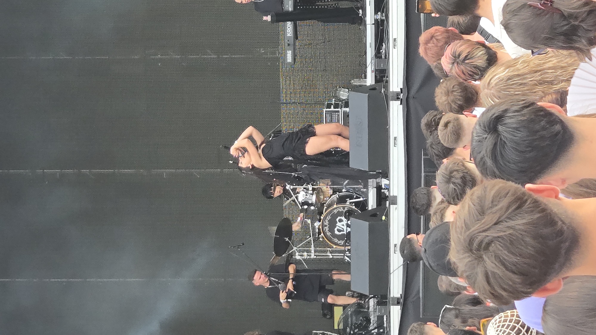 The image depicts an outdoor musical performance at the Untold Festival in Cluj-Napoca, Romania. In the foreground, the backs of an audience are visible, with various hairstyles. On the stage, a female singer in a black dress is the central focus, with her arms raised and mouth open, appearing to be singing or shouting into a microphone. To her left, a male musician in a black t-shirt and shorts is visible, holding a microphone. Behind the singer, a drummer is seated at a drum set. Further to the right, a keyboardist is playing a Korg keyboard. The stage has black speakers and a large backdrop. The overall environment suggests a large-scale music festival.