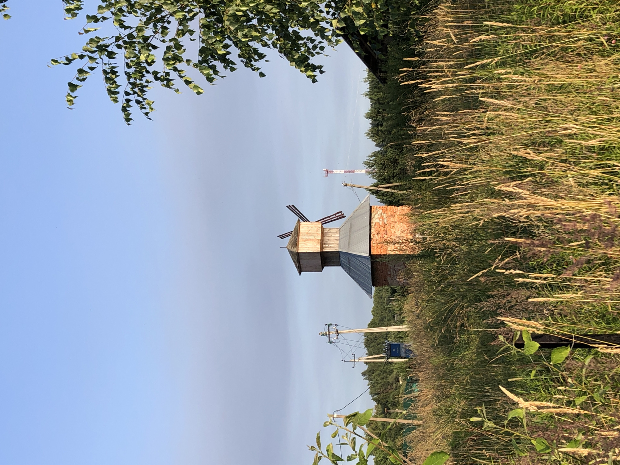 This image depicts a rural landscape in Makeikha, Russia, featuring a rustic brick structure resembling a windmill, surrounded by tall, dry grass and foliage under a clear blue sky. The windmill is made of red brick for its base and has a wooden upper section with visible red blades. In the background, several utility poles with electrical wires and a distant cell tower are present. A portion of a leafy green tree is visible on the right side of the frame, partially obscuring the sky and the background. The scene is characterized by natural lighting, suggesting it was taken during daylight hours, likely late afternoon given the long shadows cast by the vegetation. No people or animals are visible in the image.