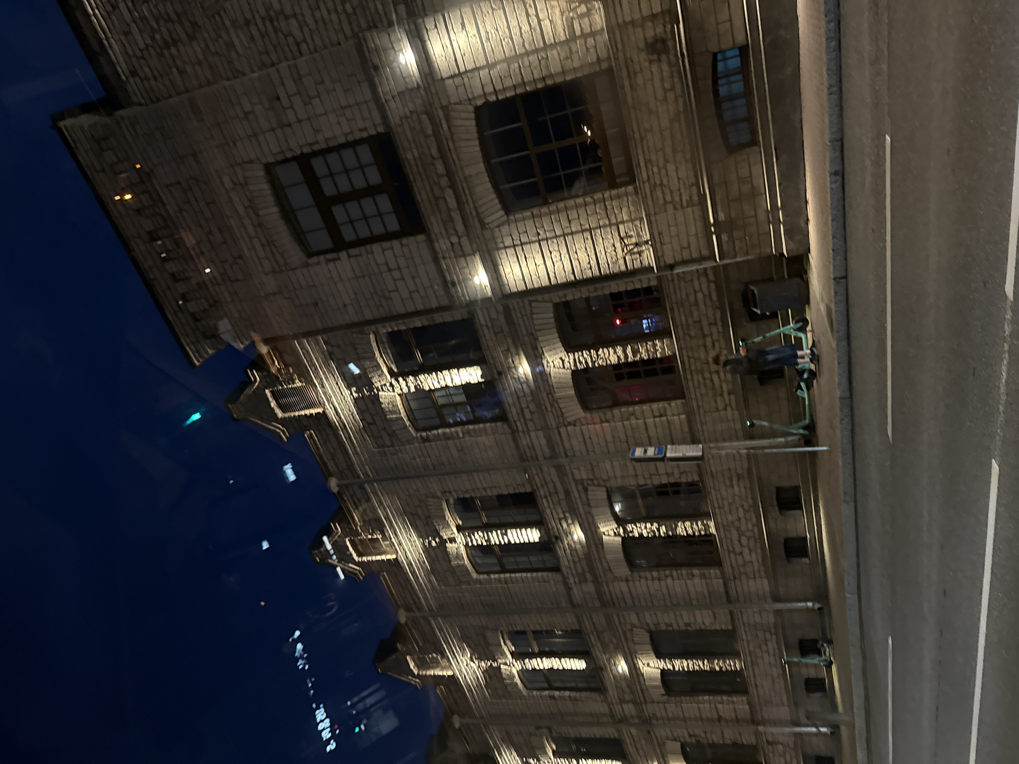 The image shows a street view of a building facade at night in Tallinn, Estonia. The building is constructed of light-colored brick and features multiple stories with numerous windows, many of which are illuminated from within. The facade is lit by external lights, creating strong shadows and highlighting the architectural details. In the foreground, a paved road with white lane markings is visible. Along the sidewalk in front of the building, several electric scooters are parked. A person is standing near one of the scooters, looking at their phone. The sky above is dark blue, indicating nighttime. The overall scene suggests an urban environment, possibly a commercial or residential area in Tallinn.
