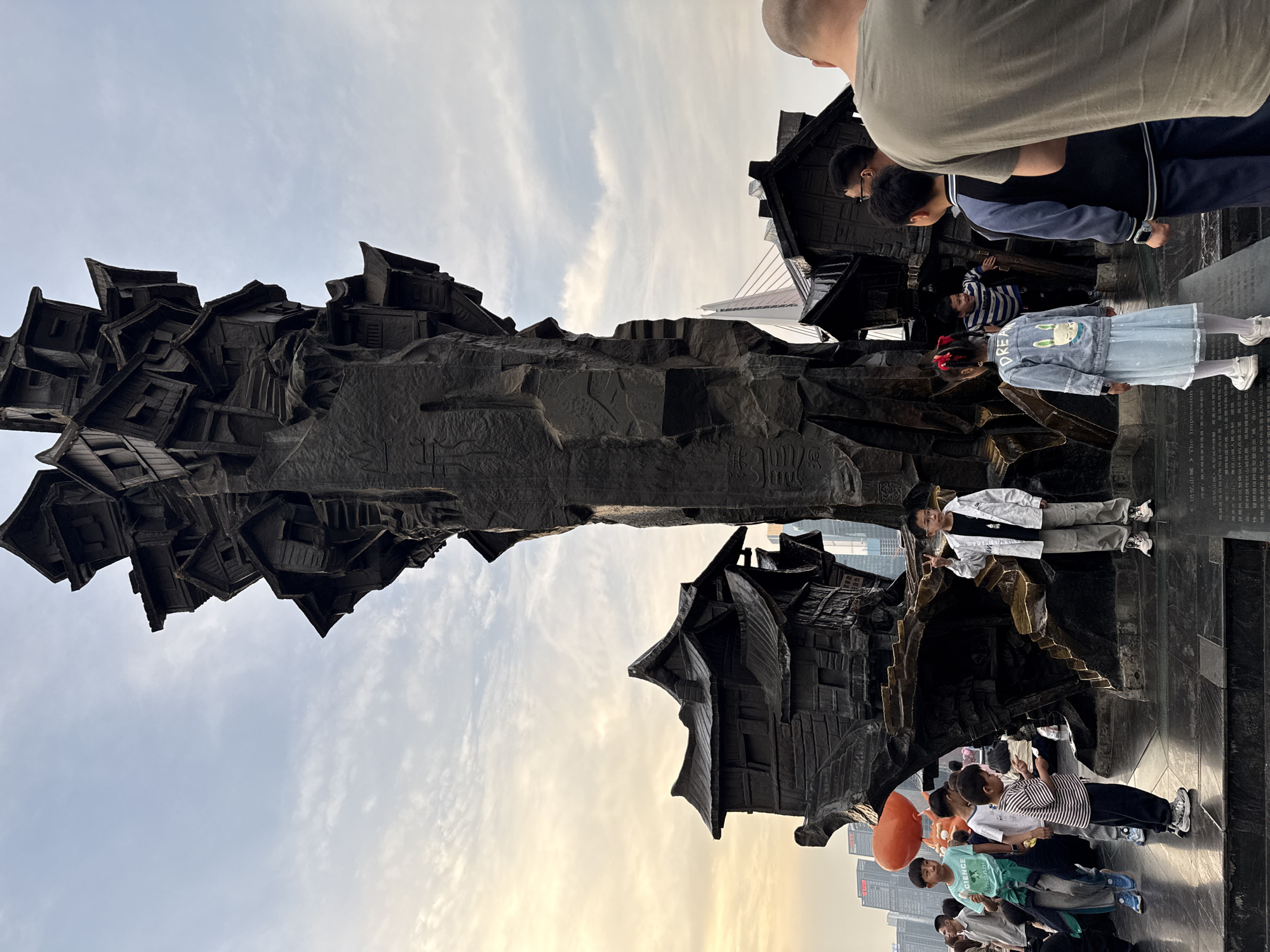 People are gathered around the Arhats in the Luohan Temple (罗汉寺) in the city of Yuzhong District, China. The artwork appears to be made of a dark metal with etchings, and is shaped like buildings. A child with an orange balloon is in the crowd. In the background, skyscrapers, a bridge, and a cloudy sky are visible.