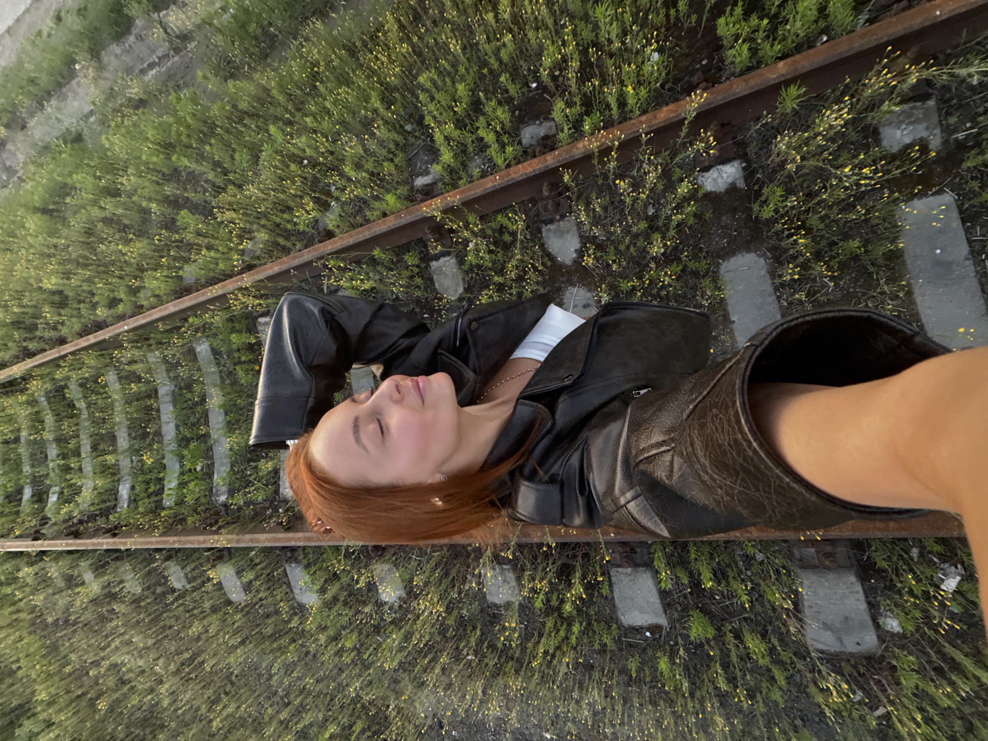 A young woman with red hair is lying on her back on abandoned train tracks, taking a selfie. She is wearing a black leather jacket and a white top. Her eyes are closed, and her head is tilted back. The train tracks are overgrown with grass and small yellow flowers. The scene appears to be located in a rural area outside of Chaikovsky, Russia.
