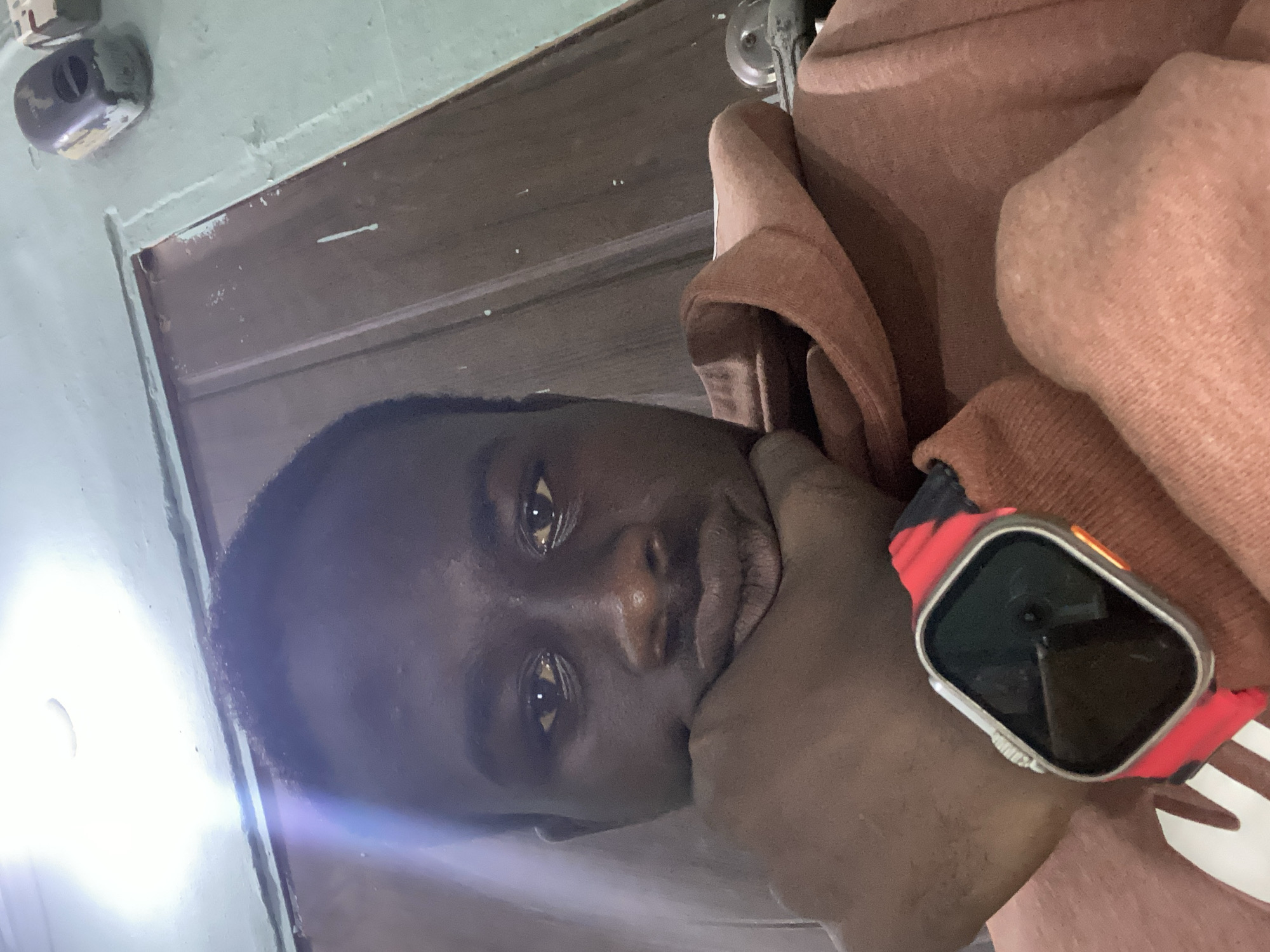 A close-up, low-angle selfie shows a young Black person with dark skin and short hair, resting their chin on their left hand. They are wearing a light brown hoodie and a silver smartwatch with a bright red strap on their left wrist. The background is a wall painted a light teal color, with a brown wooden door on the right and a bright light source at the top left of the frame. A few objects are visible on the wall, including a dark-colored electronic device and part of a beige box.