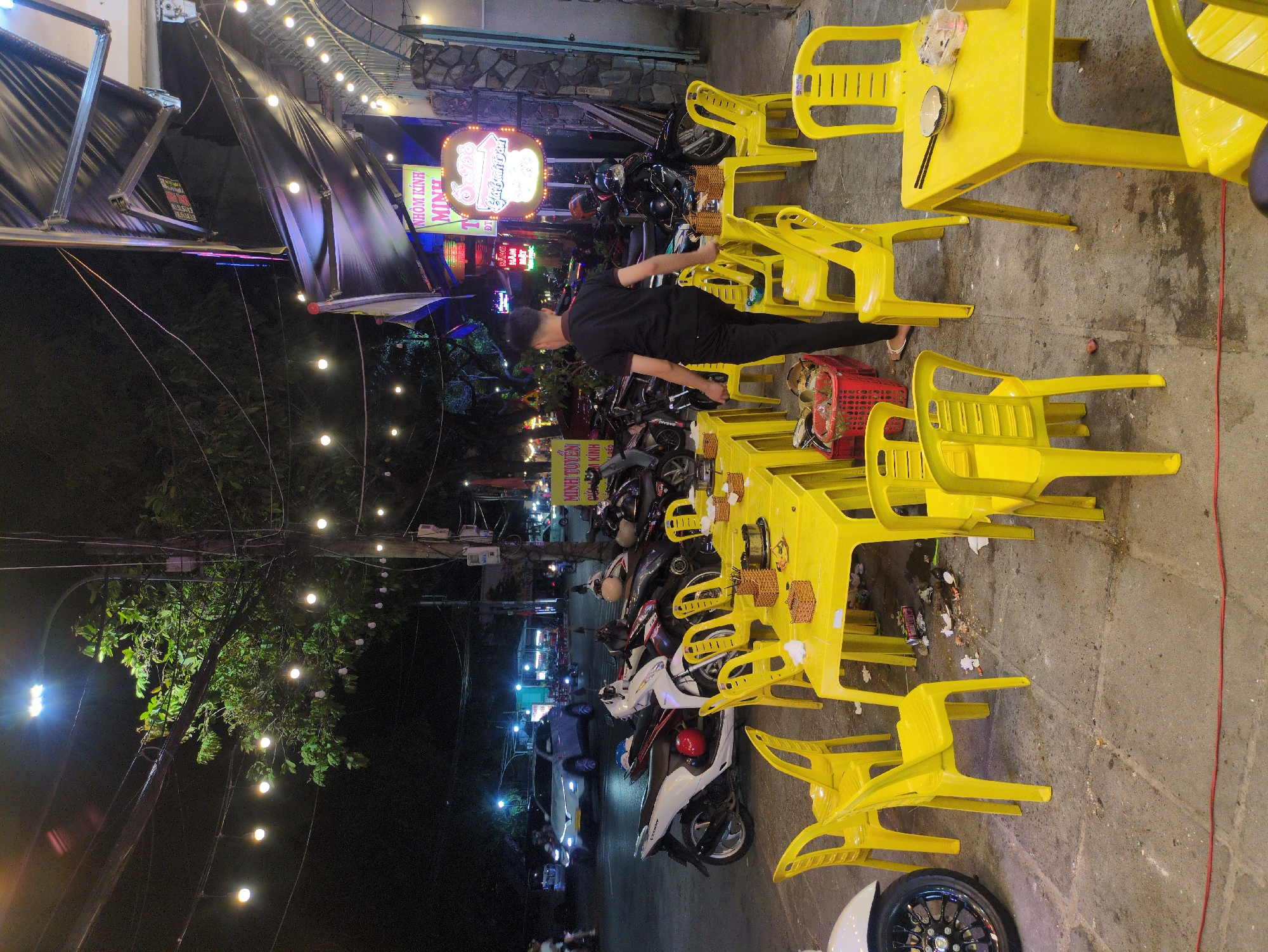 The image shows an outdoor dining area in Tuy Hòa, Vietnam, at night. String lights illuminate the scene. Multiple yellow plastic tables and chairs are arranged on the sidewalk. A person wearing black clothing stands amidst the tables, potentially attending to them. Several parked motorbikes are visible, lining the street adjacent to the dining area. A storefront with illuminated signs in Vietnamese is partially visible in the background. The pavement is concrete.