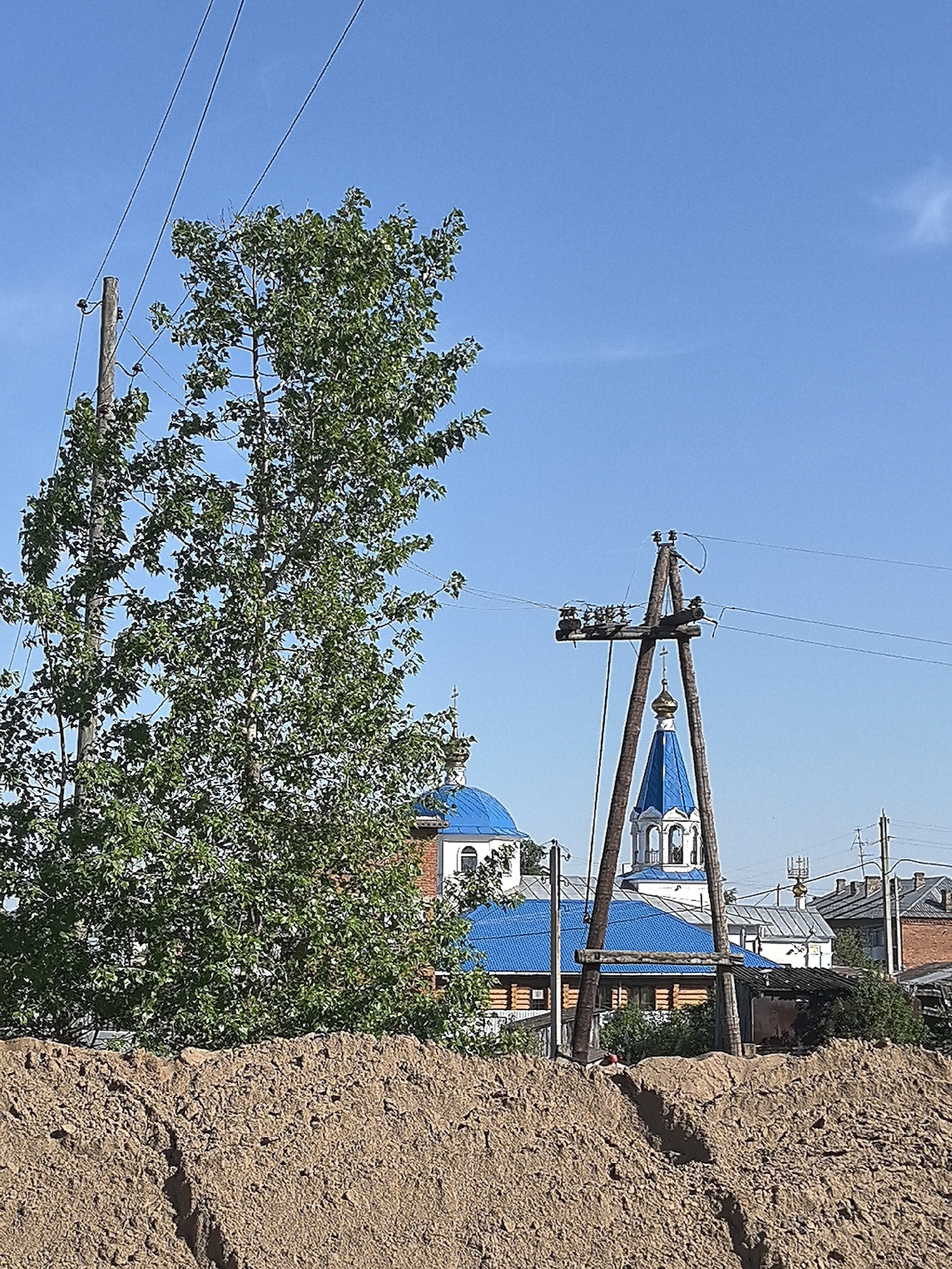 The image captures a scene in Noviy, Russia, presenting a confluence of natural and man-made elements. A mound of brown earth dominates the foreground. Beyond this, a lush green tree stands prominently to the left, partially obscuring a wooden electrical pole with power lines extending across the frame. The midground features a partially visible church with blue domes and a tower, framed by the foliage and the electrical infrastructure. Several buildings are visible in the background. The sky is a clear blue, with minimal cloud cover.