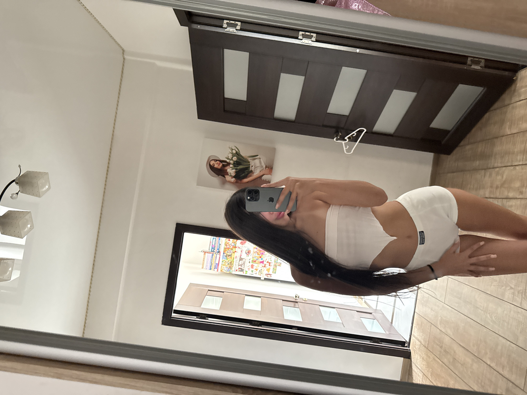 The image shows a young woman with long dark hair, taking a selfie in a mirror. She is wearing a white bandeau top and white shorts. Her right arm is raised, holding a smartphone, and her left hand is resting on her hip. The background of the image is a room with wooden floors and white walls. There is a dark brown door on the right side of the frame with frosted glass panels, and another door on the left side, partially open, revealing a hallway with colorful posters on the wall. A framed picture of a woman holding flowers hangs on the wall to the left of the dark brown door. There is a white hanger on the dark brown door. The lighting suggests an indoor environment, likely a bedroom or a dressing area. There is no discernible indication of the specific location within Chișinău, Moldova.