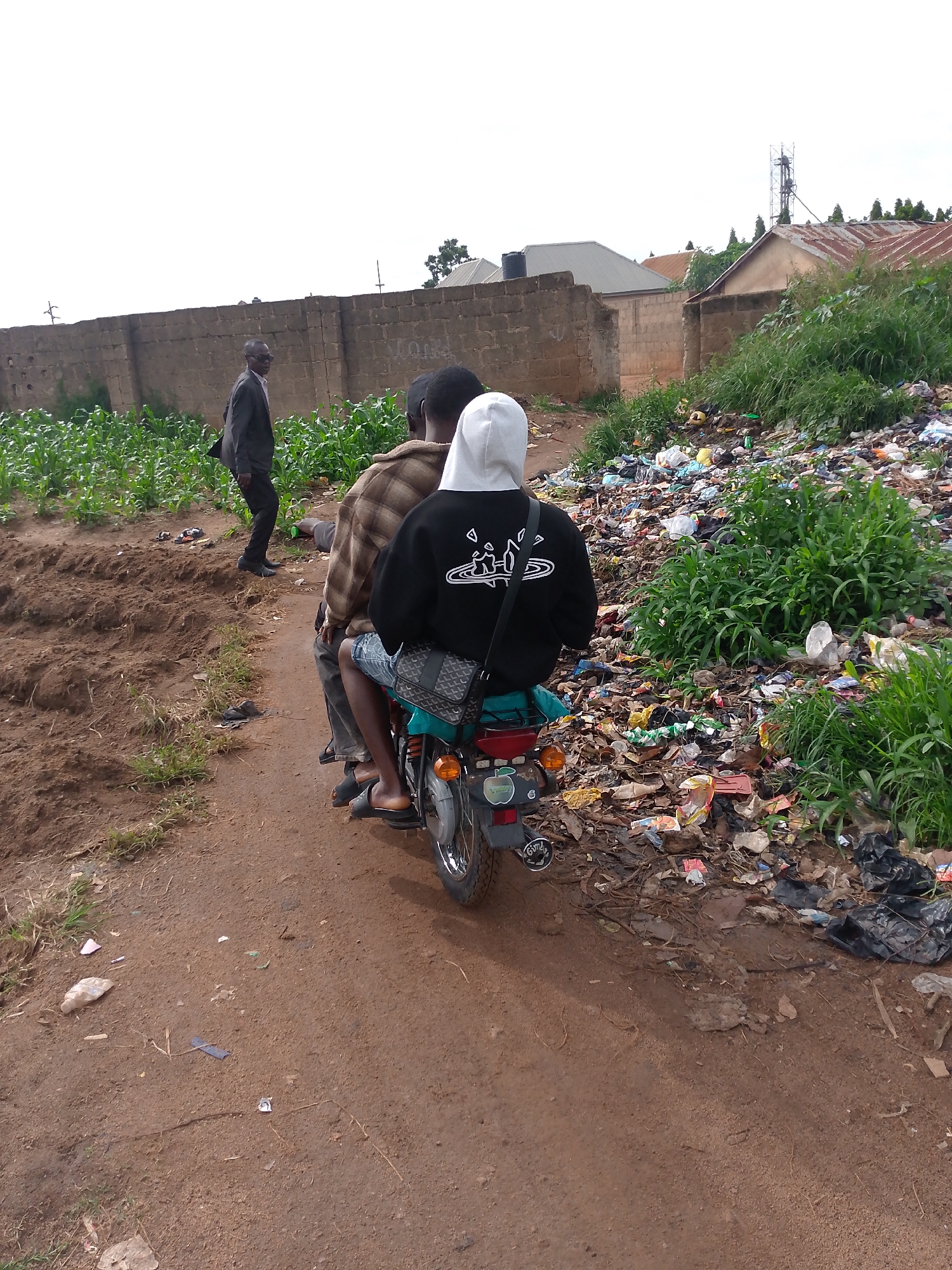 In Bukuru, Nigeria, a dirt road runs adjacent to a field of young cornstalks, bordered by a low stone wall. On the right side of the road, a large pile of refuse and assorted debris has accumulated. Three individuals are present. Two men are seated on a motorcycle, the driver wearing a plaid shirt and jeans, and the passenger behind him in a black hoodie with white detailing and a white hood, and shorts. A third man, dressed in a dark suit, walks away from the motorcycle towards the cornfield. The scene appears to be outdoors in an undeveloped area with buildings and a cell tower visible in the background.