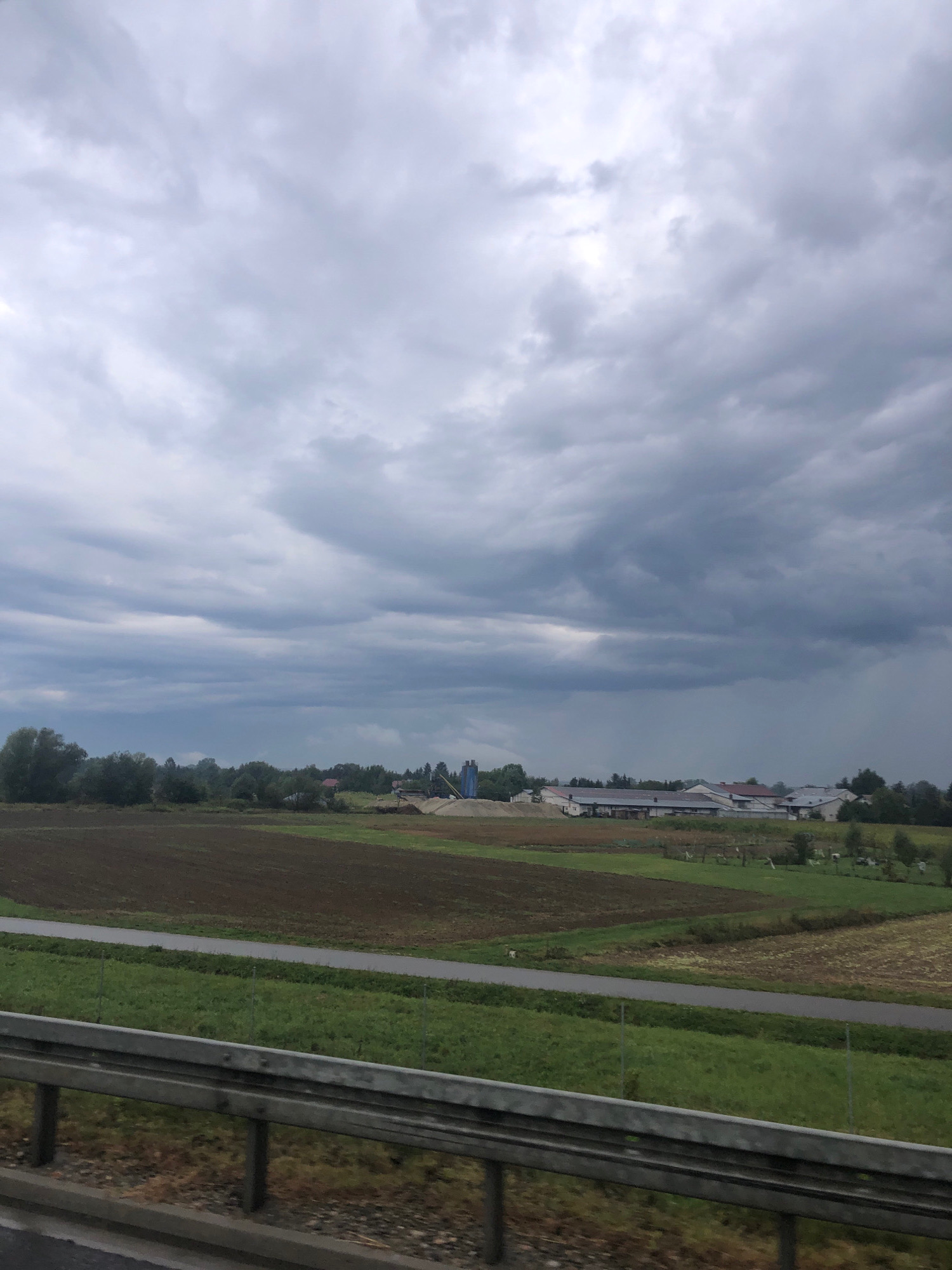 A wide-angle, eye-level shot captures a rural landscape under a heavily overcast, gray sky. The scene appears to be a farming area in or near Smolarzyny, Poland. In the foreground, a metal guardrail runs horizontally across the bottom of the image, with a paved surface and a grassy verge behind it. Beyond the verge, cultivated fields dominate the midground. Some fields show dark brown, recently tilled soil, while others are covered in green grass or have lighter brown, stubbled crops. A narrow, gray asphalt road cuts through these fields. In the distance, a cluster of agricultural buildings with white walls and dark roofs forms a small settlement. Adjacent to the buildings, a large pile of light-colored material, possibly gravel or sand, is visible, along with what appears to be construction or industrial equipment. A line of trees forms a dark green border behind the buildings, separating them from the sky. The sky itself is filled with thick, ominous gray clouds, suggesting an impending rain shower or a generally gloomy day. There are no people or animals visible in the scene. The overall impression is one of a working agricultural landscape on a somber day.