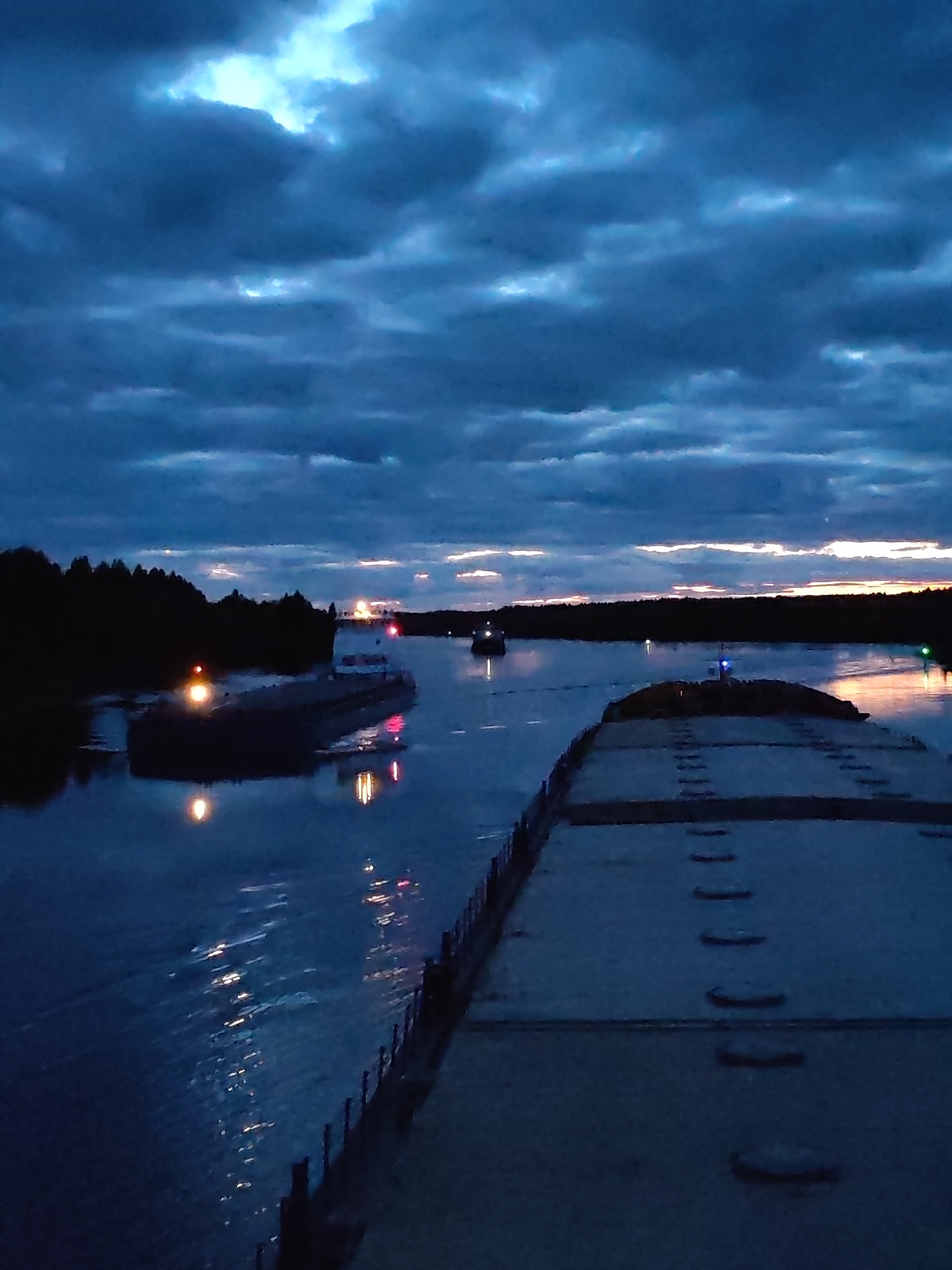 The image shows a night scene on a river, possibly the Volga River, with several barges or ships navigating the waterway. In the foreground is the deck of a barge with visible hatch covers. Two other vessels are visible in the middle ground, illuminated by their navigation lights, reflecting on the water's surface. The riverbanks are lined with trees, and the sky is filled with dark, cloudy formations, hinting at twilight or night. The bright area on the horizon may suggest the remnants of sunset or the beginning of dawn. The location cannot be verified as Uzhla, Russia.