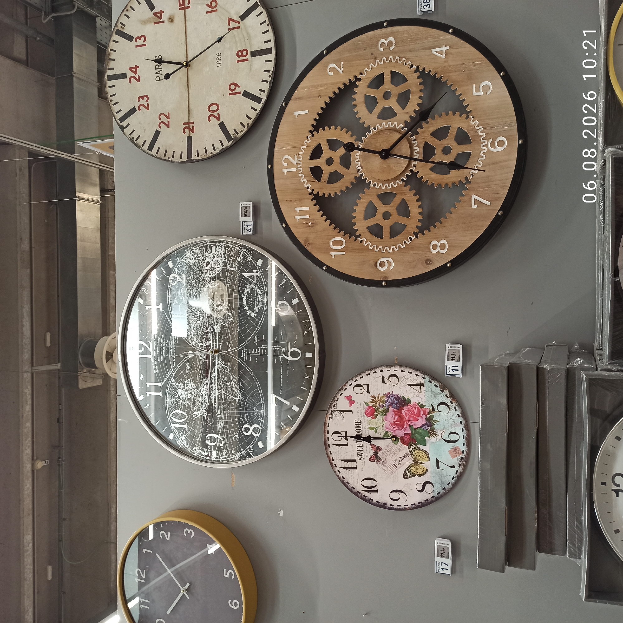 This image depicts a wall display of various clocks in what appears to be a retail store. The background is a plain gray wall. On the wall, there are at least four distinct clocks visible.
The top left features a round, dark gray clock with a yellow bezel. Its face has white hour markers and hands.
To its right, a large, round black clock with a white face displays a map of the world. It has white Arabic numerals and minute markers.
Below and slightly to the left of the world map clock is a smaller, round clock with a decorative face featuring flowers and butterflies. It has black Arabic numerals.
To the right of the decorative clock and below the world map clock is a large, round wooden clock with visible gear mechanisms. It has white Arabic numerals and black hands.
Several price tags are visible on the wall, indicating that these are merchandise for sale. One tag reads "Posizione 41 92,50". Another reads "Posizione 17 11,50". A third tag is partially visible, reading "Posizione 38".
In the foreground, at the bottom of the image, stacks of items wrapped in plastic are visible, likely also merchandise.
The lighting suggests an indoor environment, possibly a supermarket or home goods store. The overall impression is that of a retail display area.
The timestamp "06.08.2026 10:21" is superimposed on the bottom right of the image, indicating the date and time the photograph was taken. The location of Bari, Italy is not deducible from the image content itself.