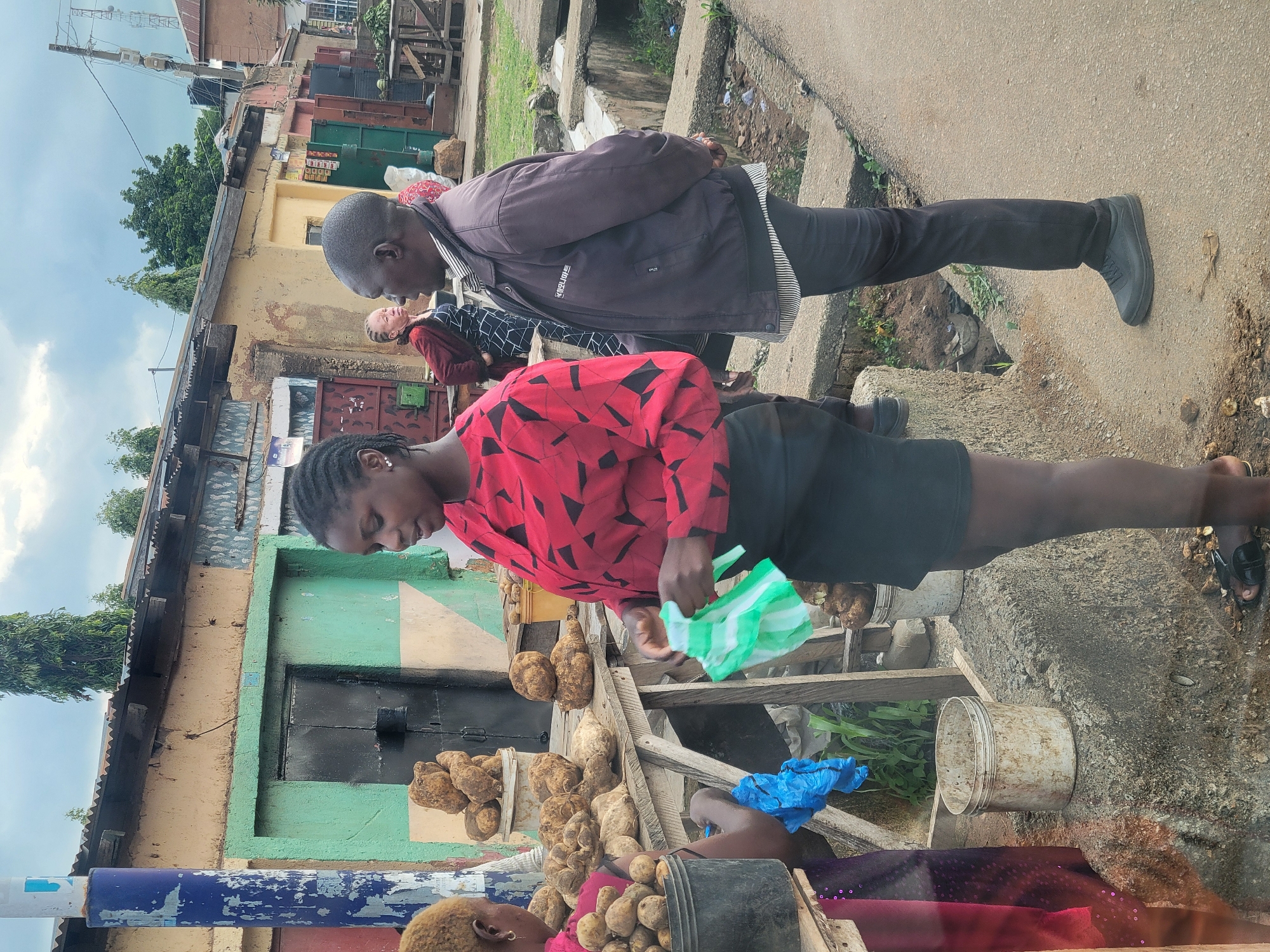 In the city of Jos, Nigeria, an outdoor market scene unfolds. A woman, wearing a red and black patterned long-sleeved shirt and a black skirt, stands in the foreground, holding a green and white striped plastic bag. She appears to be interacting with another person, whose back is mostly turned to the camera, wearing a dark jacket over a striped shirt and dark pants. Various root vegetables, likely yams, are piled on a wooden table to their left, with some in buckets. A young woman with light-colored hair is partially visible to the far left, also near the vegetables, holding a blue plastic bag. In the background, market stalls and buildings with corrugated iron roofs are visible, along with a paved road and drainage channels. The sky is overcast, suggesting a cloudy day.