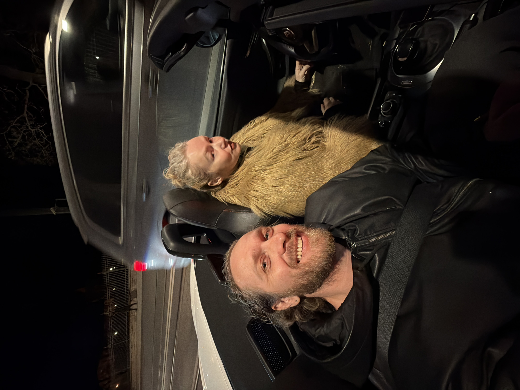 Two individuals are in what appears to be the interior of a convertible car, possibly at night. The car may be parked, given the surrounding darkness. One individual, a man, is in the foreground wearing a black jacket and a seatbelt, smiling towards the camera. The second person, a woman, is behind him, wearing a large, fluffy brown coat or jacket. The car's interior is visible with controls and seats. Based solely on the image content, the location within St. Petersburg, Russia, cannot be determined.