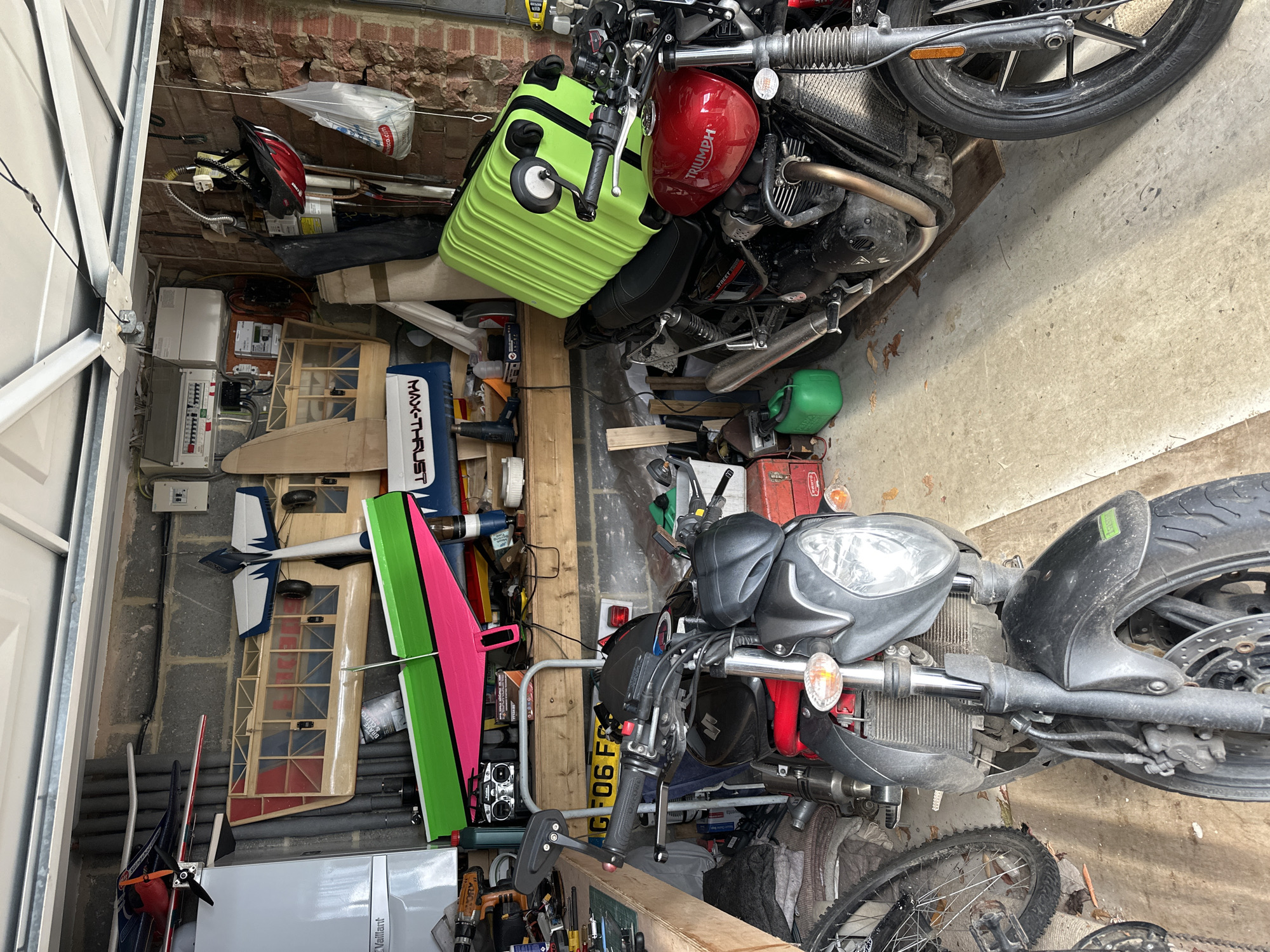 The image depicts the interior of a cluttered garage or workshop located in the Wealden District, United Kingdom. The ceiling is composed of white, paneled garage door sections. The walls consist of exposed cinder blocks on the left and rear, and exposed, somewhat distressed red brick on the right. The floor is concrete, with a section of cardboard or similar material visible under the front wheel of the left motorcycle.
No individuals are present.
On the left side, a white Vaillant boiler unit is mounted on the cinder block wall, with grey corrugated piping running horizontally above it. Above a wooden workbench along the rear wall, multiple electrical consumer units are affixed. The workbench supports various tools and objects, including drills, bottles, a radio-controlled transmitter, and a "MAX-THRUST" branded box. Several model aircraft are stored or displayed; notable examples include a bright pink and neon green model airplane positioned prominently, a wooden-framed model with red and blue accents, and a smaller white and blue model.
On the right side, a red bicycle helmet hangs from the brick wall, below which a white plastic bag, partially branded "Tesco," is suspended.
Two motorcycles are parked facing the viewer. The left motorcycle is a dark grey/black Suzuki, with its front wheel, disc brake, headlight, and handlebars clearly visible. A partial license plate, "G F06 FS," is visible behind its handlebars. To the immediate left of this motorcycle, a bicycle wheel is partially visible. The right motorcycle is a red Triumph Street Twin, identifiable by its fuel tank with the "Triumph" logo and "Street Twin" lettering on its side. A neon green hardshell suitcase with black wheels and handles rests horizontally on the rear seat of the Triumph. A small, bright green jerry can is on the floor near the rear wheel of the Triumph.