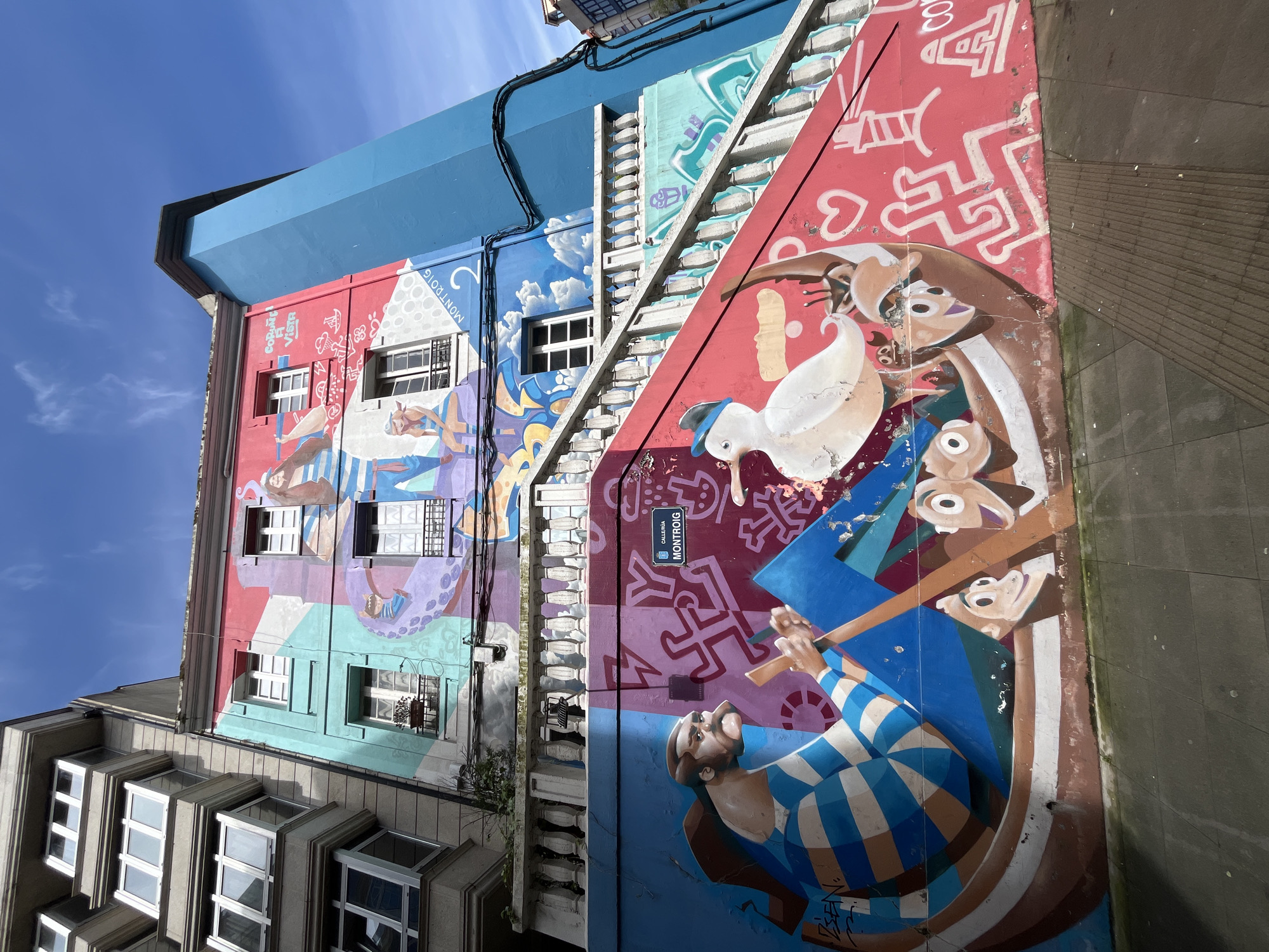 The image shows a tall building in A Coruña, Spain, adorned with vibrant, colorful murals. The murals depict a variety of figures and scenes, including what appears to be a man rowing a boat with animal figures, as well as geometric shapes and abstract patterns. The building has windows of various sizes and architectural styles, and a street sign that says "CALLE MONTROIG". The sky is blue with some clouds visible. The perspective is from a low angle, looking upwards at the building.