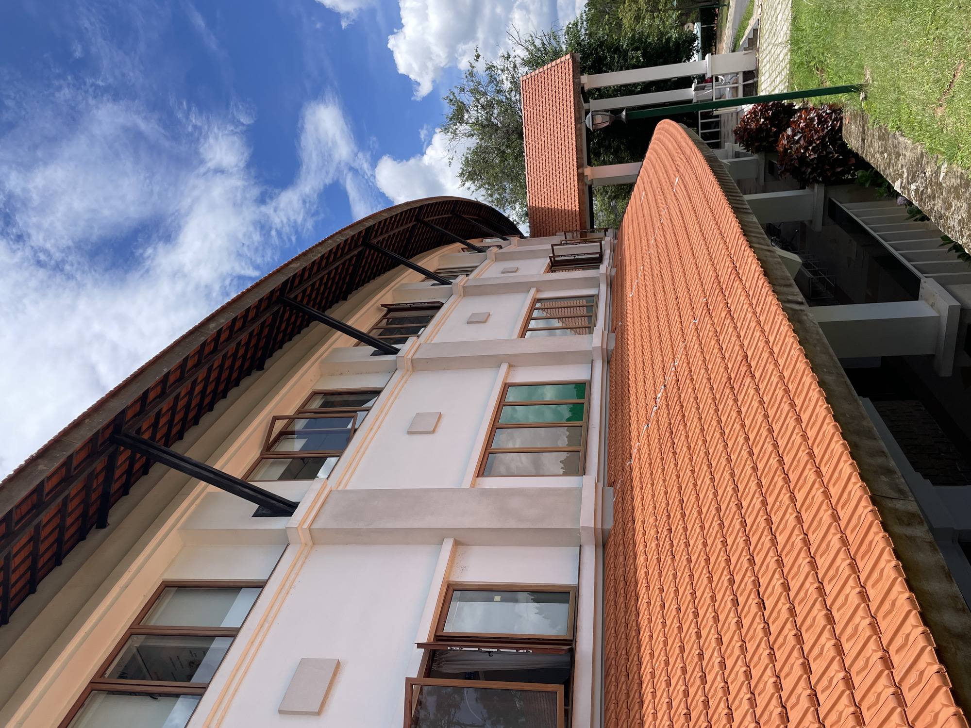 The image shows a white building with a curved roof tiled in orange terracotta. The building has several windows, some of which are open, revealing the interior. The roof is supported by dark metal beams. In the foreground, a wide expanse of the orange tiled roof slopes downwards. Behind the building, lush green trees and a bright blue sky with scattered white clouds are visible. The architectural style appears to be modern with some traditional elements. The location is specified as the Dinh 2 building, a prominent tourist attraction in Da Lat, Vietnam, known for its distinctive architecture and beautiful surroundings. There are no people visible in the image. The overall scene is one of a well-maintained building set within a natural, verdant environment.
