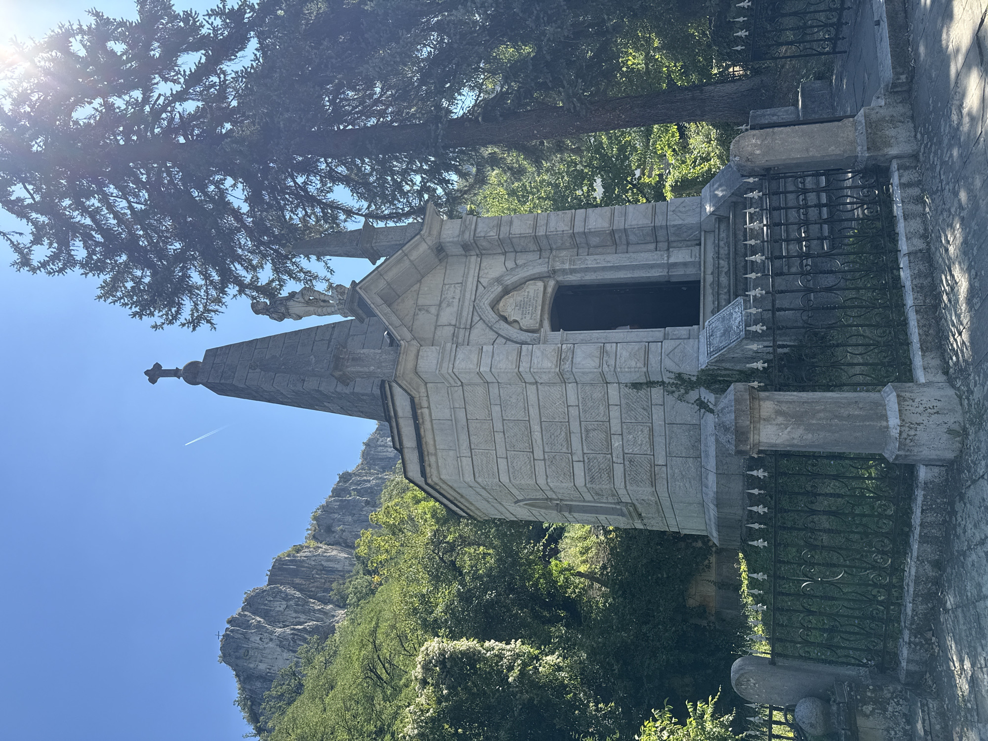 The image depicts a memorial structure with a fence in front of it, located in Dryanovo, Bulgaria. The memorial is a stone building with a pointed roof topped by a cross. A statue of a person stands on the roof near the front. The building has an entrance with a dark door and steps leading up to it. The fence is made of metal with decorative elements. Behind the memorial, there are trees and a rocky mountain. The sky is clear and blue, with a contrail from an airplane visible. The scene appears to be a historical or religious site, possibly a cemetery or a monument.