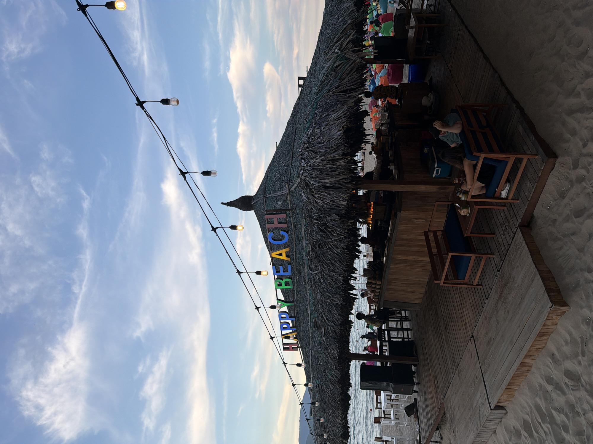The image shows a beach bar with a thatched roof and colorful "HAPPY BEACH" signage. The bar is set up on a wooden platform on a sandy beach. There are people sitting at tables and on beanbag chairs, some are relaxing while others are standing or walking around. The sea is visible in the background, with mountains in the distance. String lights are hanging over the bar area. The scene appears to be in Nha Trang Ward, Vietnam, judging by the presence of beach bars and the overall ambiance.