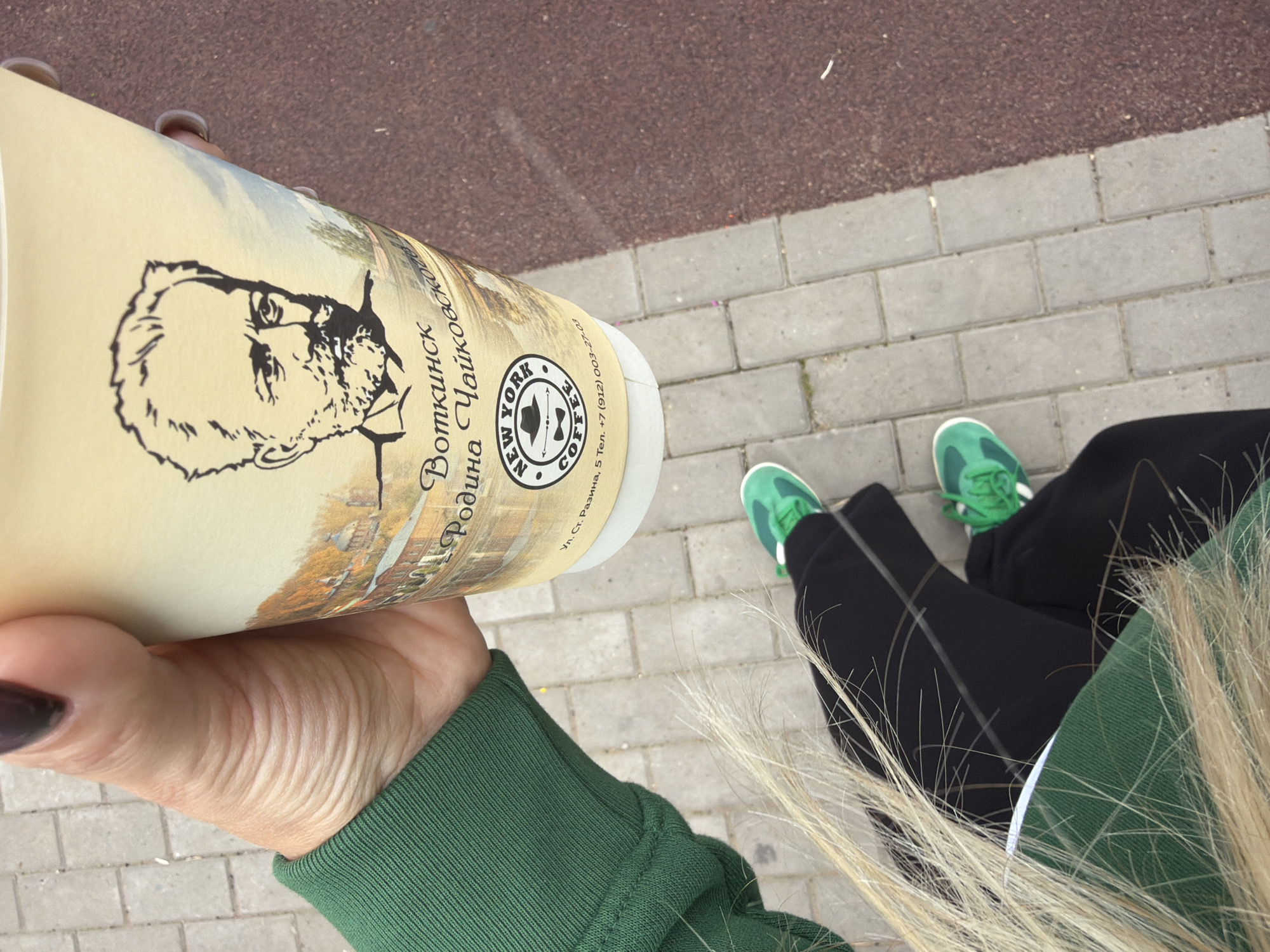 This media file shows a person holding a disposable coffee cup outdoors. The cup features a portrait of a man and text in Russian, identifying it as from "Votkinsk, the Homeland of Tchaikovsky," with a New York Coffee Company logo and contact information. The person is wearing a green hoodie and black pants. Their legs, clad in bright green sneakers, are visible, suggesting they are sitting or crouching. The background consists of a textured, reddish-brown surface transitioning to a paved area with grey rectangular bricks. The lighting suggests it is daytime, possibly overcast given the lack of harsh shadows. The overall scene appears casual and relaxed, possibly during a break or outing in an urban or park-like setting.
