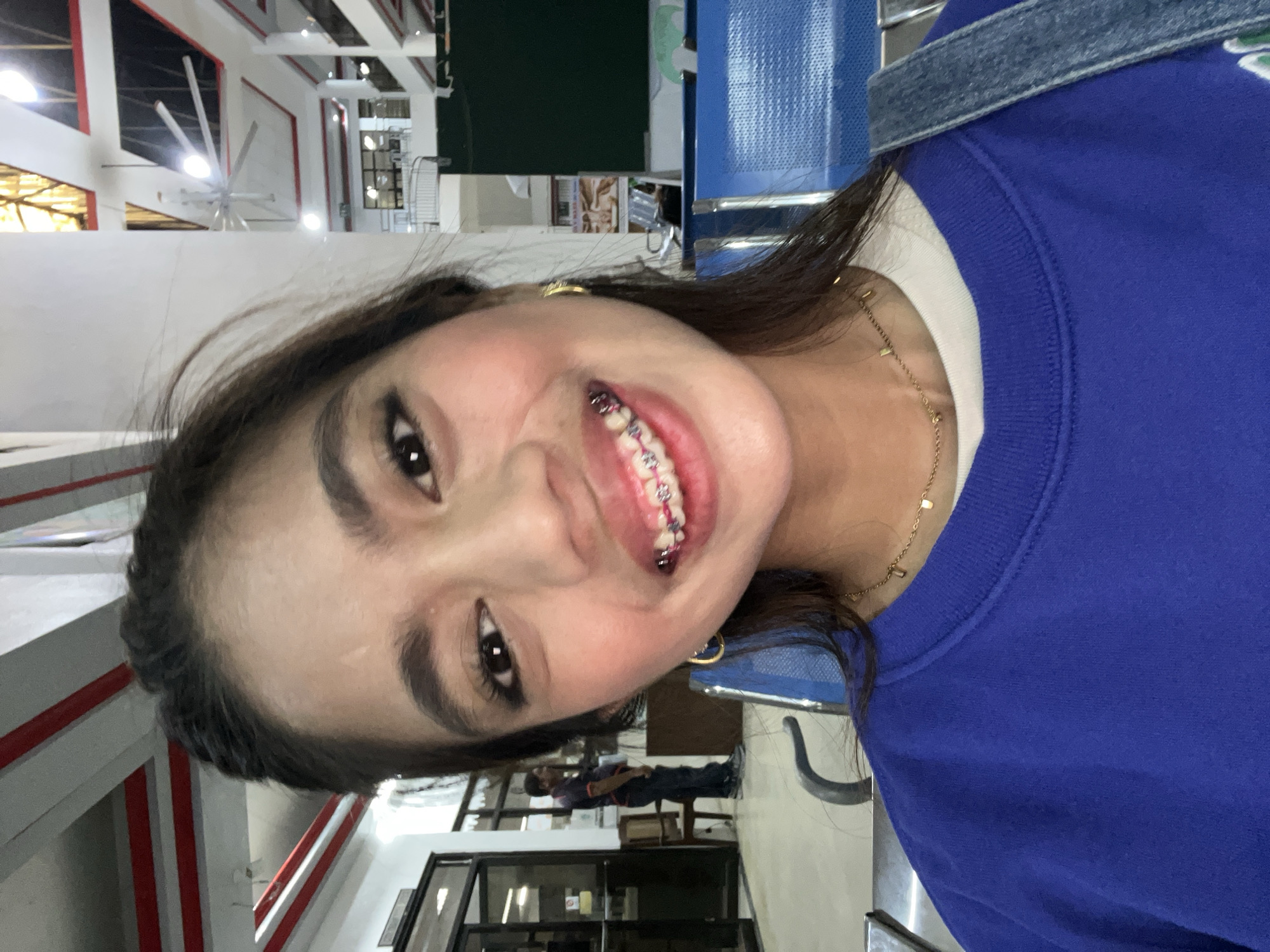 A young woman with dark hair, wearing a blue sweatshirt and a gold necklace, smiles at the camera. She has braces on her teeth, and gold hoop earrings. The background appears to be a waiting area with blue chairs and a man standing in the distance. The ceiling has exposed beams and a large industrial fan. This location is likely the Panabo City Hall, a municipal building in Panabo City, Davao del Norte, Philippines.