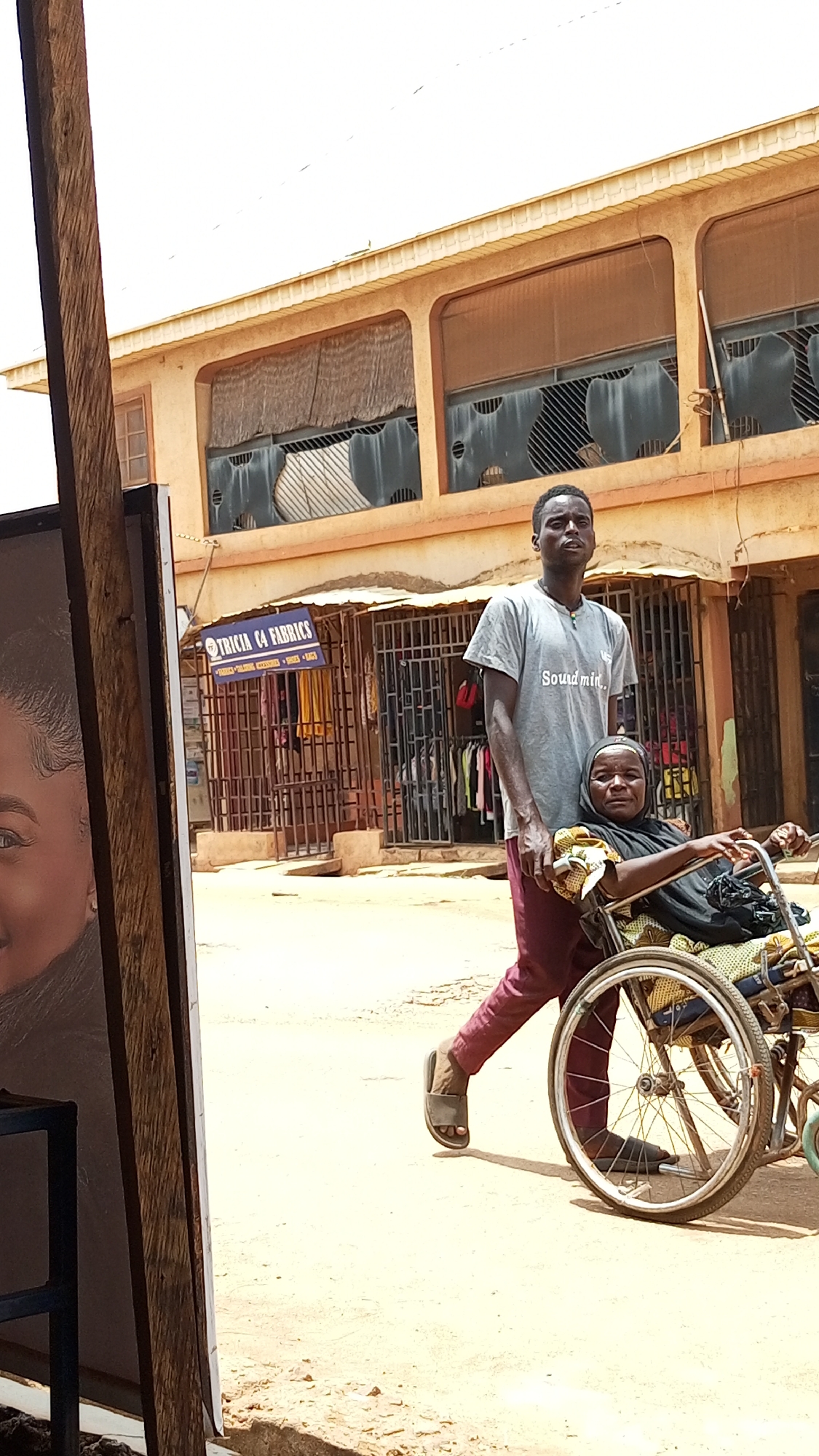 In Sabon Tasha, Nigeria, a young man in a grey t-shirt and maroon pants pushes a woman in a wheelchair down a dirt road. The woman wears a black hijab. They are in front of a building with a sign that reads "TRICIA CA FABRICS." The building has metal gates in front of the entrance, and clothes are hanging inside. A poster obscures the left side of the image.
