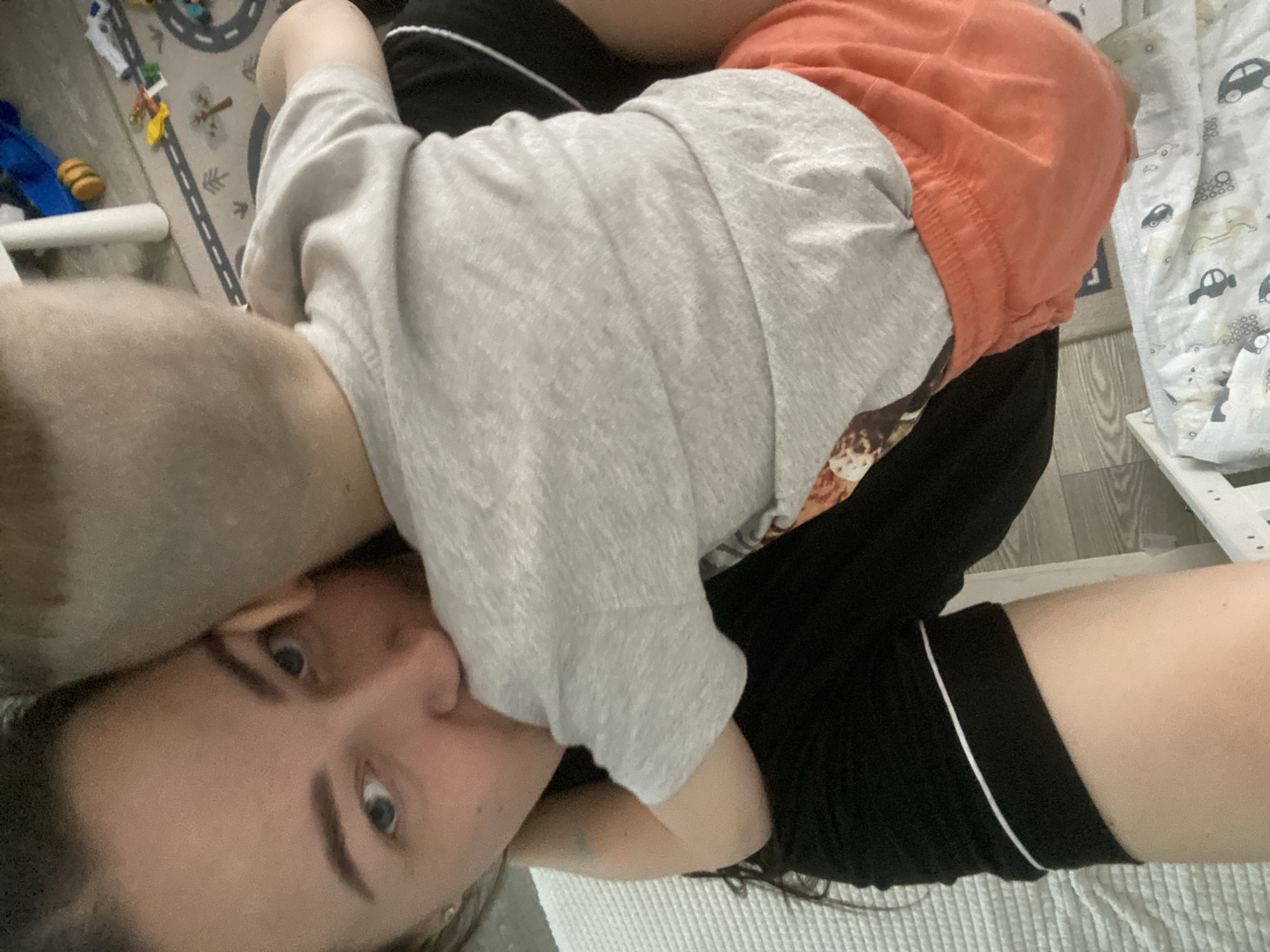 A woman with blue eyes and fair skin is holding a child. The child is wearing a gray shirt and orange shorts. The woman is wearing black shorts with a white stripe. They are on a patterned white blanket and the child appears to be resting. There is a patterned rug and a child's play area in the background. The location is not discernible from the image.