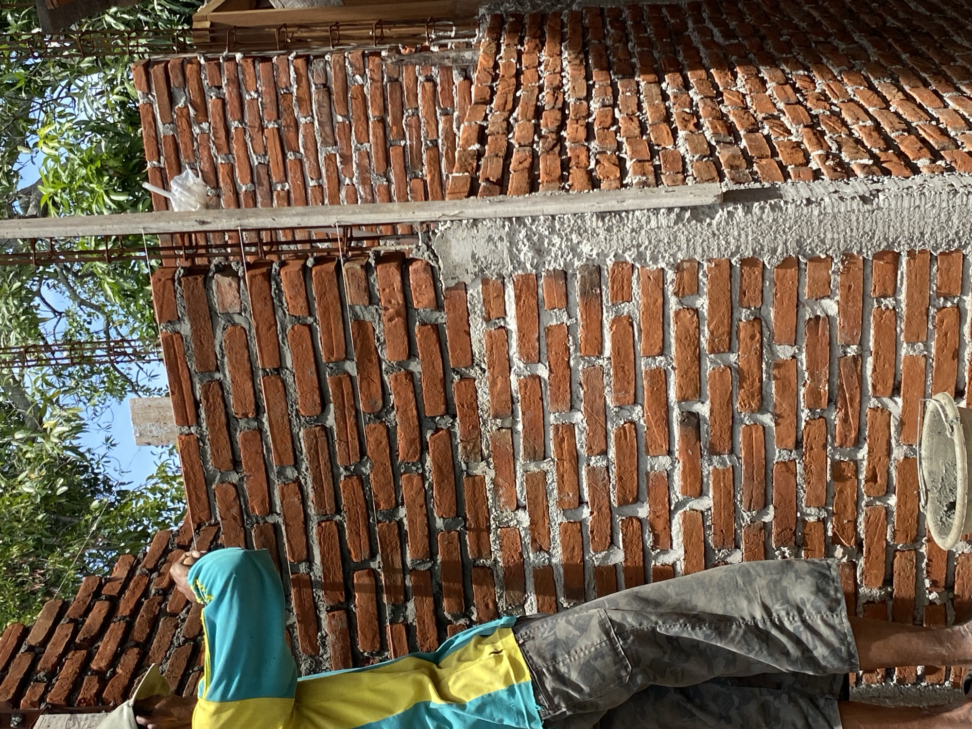 A person wearing a light green cap, a yellow and turquoise long-sleeved shirt, and camouflage pants is working on building a brick wall. The person is standing on the left side of the frame, facing towards the wall and appears to be placing or adjusting bricks. The wall is made of red bricks and mortar. There is scaffolding made of metal bars and wooden planks visible behind the wall, suggesting ongoing construction. Green foliage and a blue sky are visible in the background. A concrete pillar is positioned vertically in front of the wall on the right side. A bucket filled with a grey substance, likely mortar, is on the ground at the bottom of the frame. The scene appears to be an outdoor construction site, possibly in Banda Aceh, Indonesia, given the foliage and architectural style which are common in tropical regions of Southeast Asia.