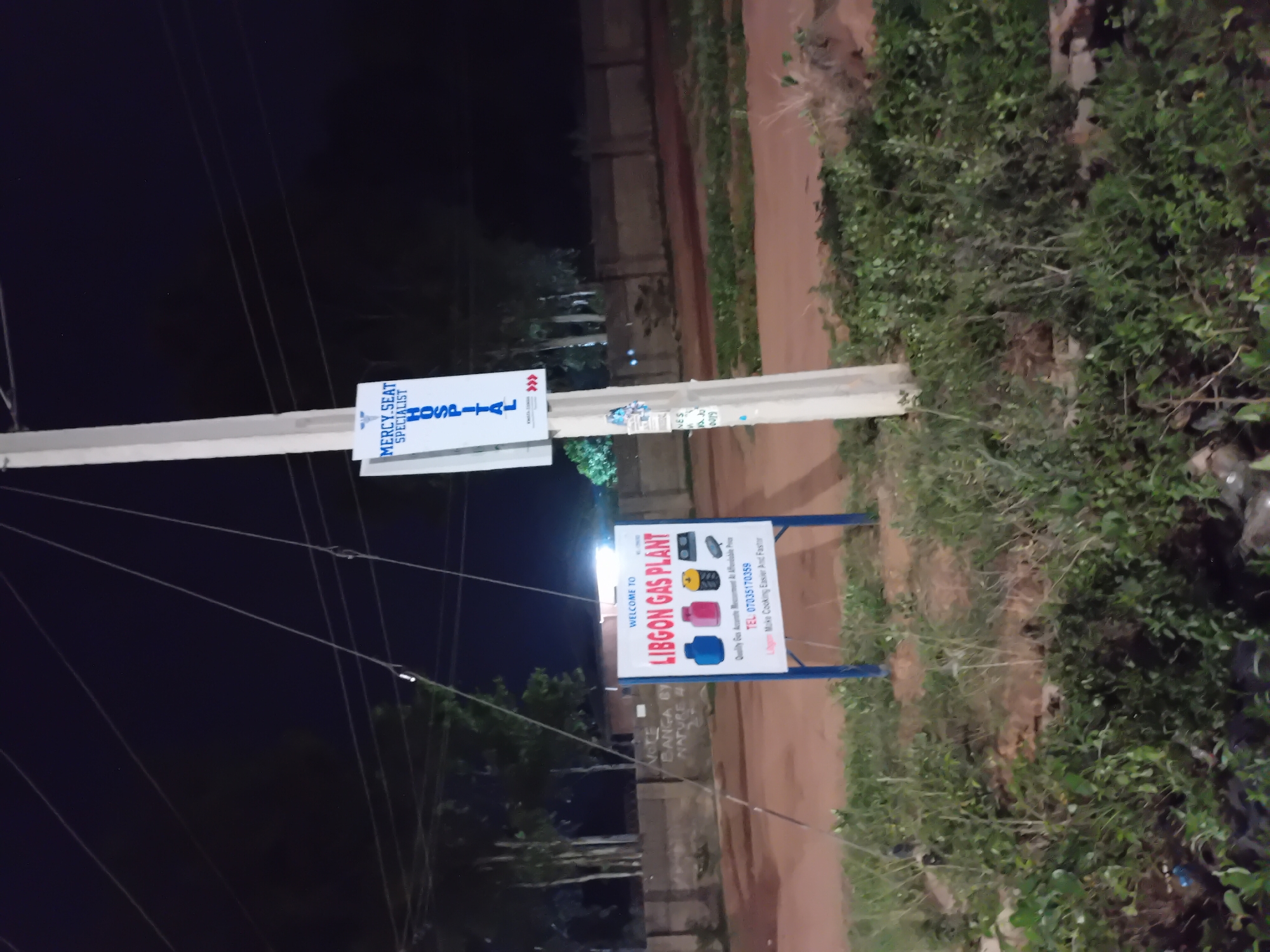 The image shows a pole in Barakin Angundi, Nigeria, with two signs attached. The top sign advertises Mercy Seat Specialist Hospital. The lower sign reads "Welcome to Libgon Gas Plant" and includes a phone number, 07035170359. Vegetation is visible on the right side of the image. It is nighttime.