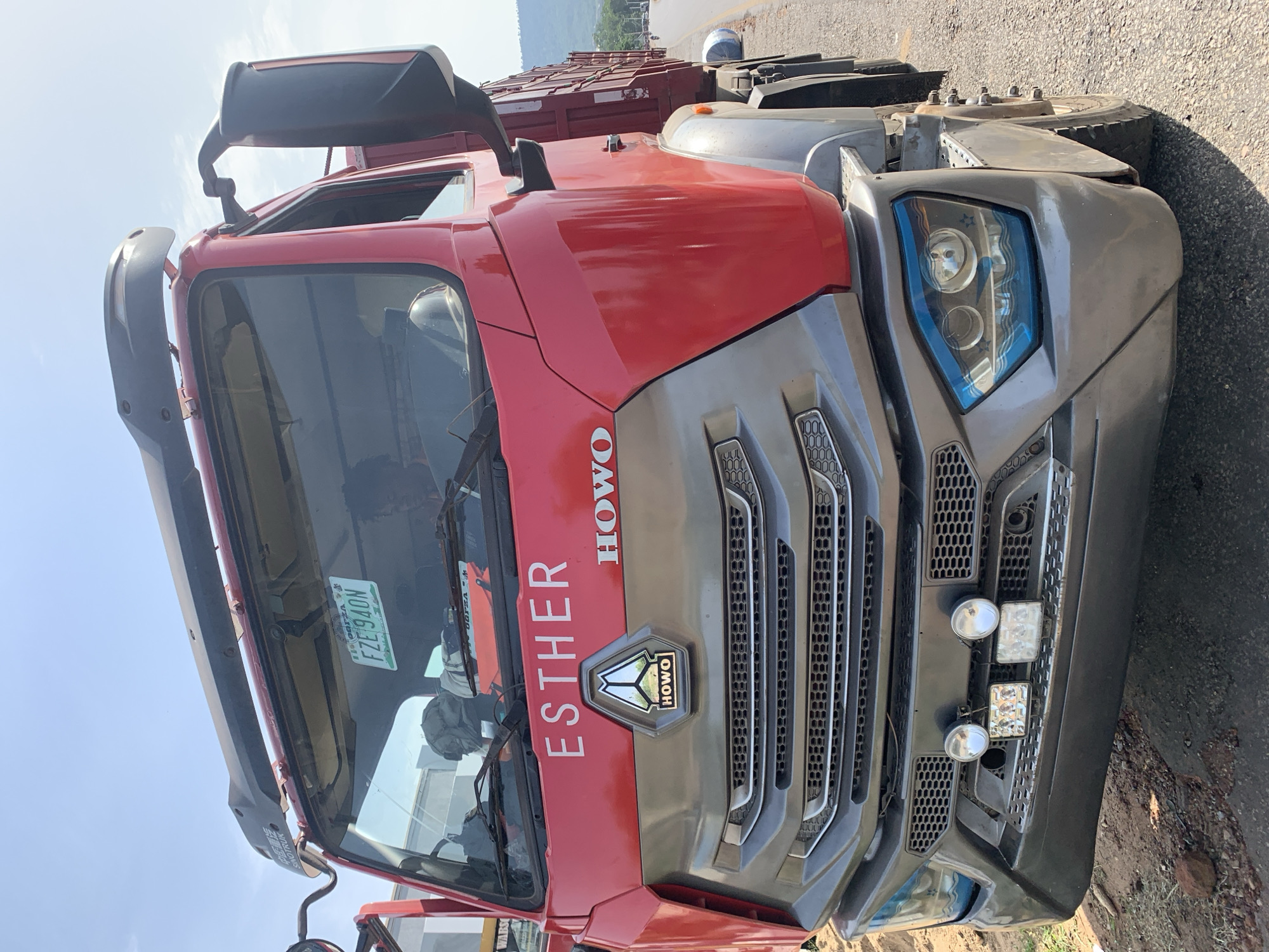 A close-up, rotated shot of the front of a red and grey Howo truck with the name "Esther" emblazoned in white letters on the red cab. The truck is parked on the shoulder of a road in what appears to be an outdoor, rural or semi-rural setting. The truck’s grill features the Howo logo. A Nigerian license plate with "FZE 940N" is visible through the windshield. The exact location is Zango-ndaji, Nigeria.