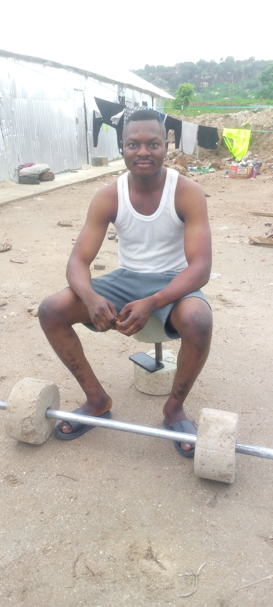In Indo, Nigeria, a young man sits outdoors in a gritty, unpaved area next to a makeshift weightlifting setup. He wears a white tank top and grey shorts, and black flip-flops. He holds his hands clasped in his lap. A barbell, constructed with a metal bar and two large concrete discs as weights, rests on the ground in front of him. To his right, a smartphone sits on a concrete block, one of two that appear to be used as weight stands. Behind the man, a corrugated metal structure, possibly a dwelling or workshop, is visible, with laundry hanging on a line in front of it. The environment suggests a rural or impoverished urban setting, characterized by dirt, rocks, and scattered debris. The sky is overcast.