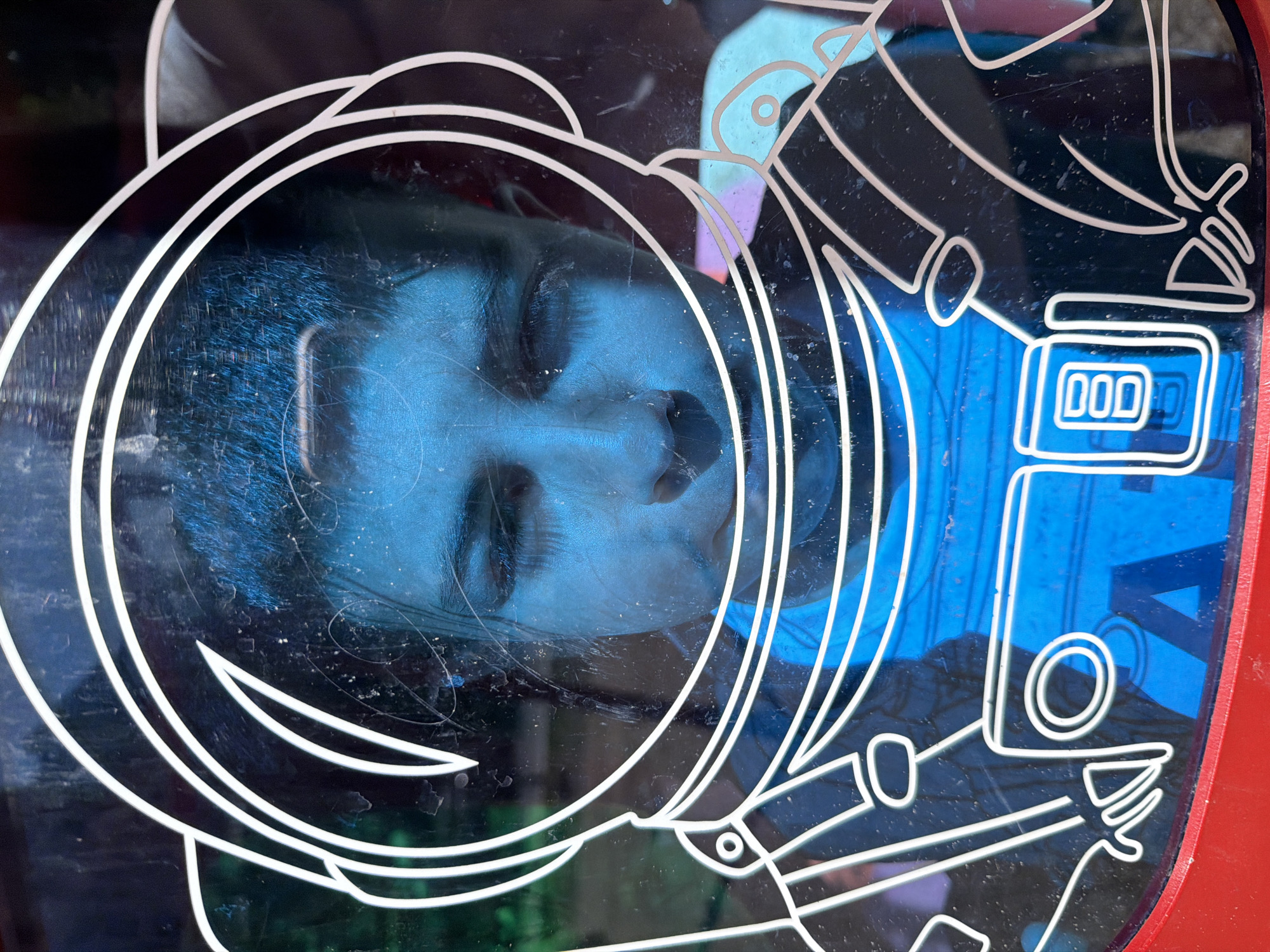The image shows a reflection of a person's face inside an astronaut helmet design. The helmet is outlined in white on a dark background, likely a window or a display. The person has their eyes closed, and their face is tinted blue, suggesting an artificial light source. The astronaut suit is rendered in a simplistic, cartoon-like style, also outlined in white. The background visible through the display appears to contain blue and red elements. The location, Yerevan, Armenia is not evident from the image itself, but is only possible to know from the image's metadata.