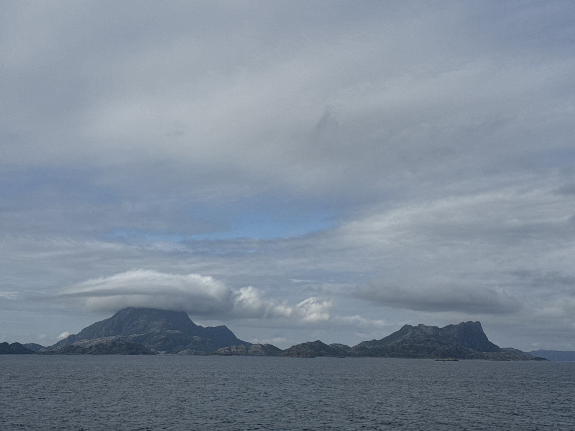 The image depicts a vast expanse of water in the foreground, with a mountainous archipelago rising from the sea in the background. The sky is overcast with a mix of gray and white clouds, with a few patches of blue sky visible. The mountains are rugged and rocky, with sparse vegetation. One prominent peak on the left is partially obscured by a lenticular cloud formation. The water is a deep blue-gray, with gentle waves. The scene appears to be a coastal landscape, likely in a northern region given the cloudy sky and rugged terrain. The location is identified as Stokkvågen, Norway, a coastal village known for its dramatic scenery and proximity to the sea. There are no people or man-made structures clearly visible in this particular shot, emphasizing the natural and remote beauty of the location.